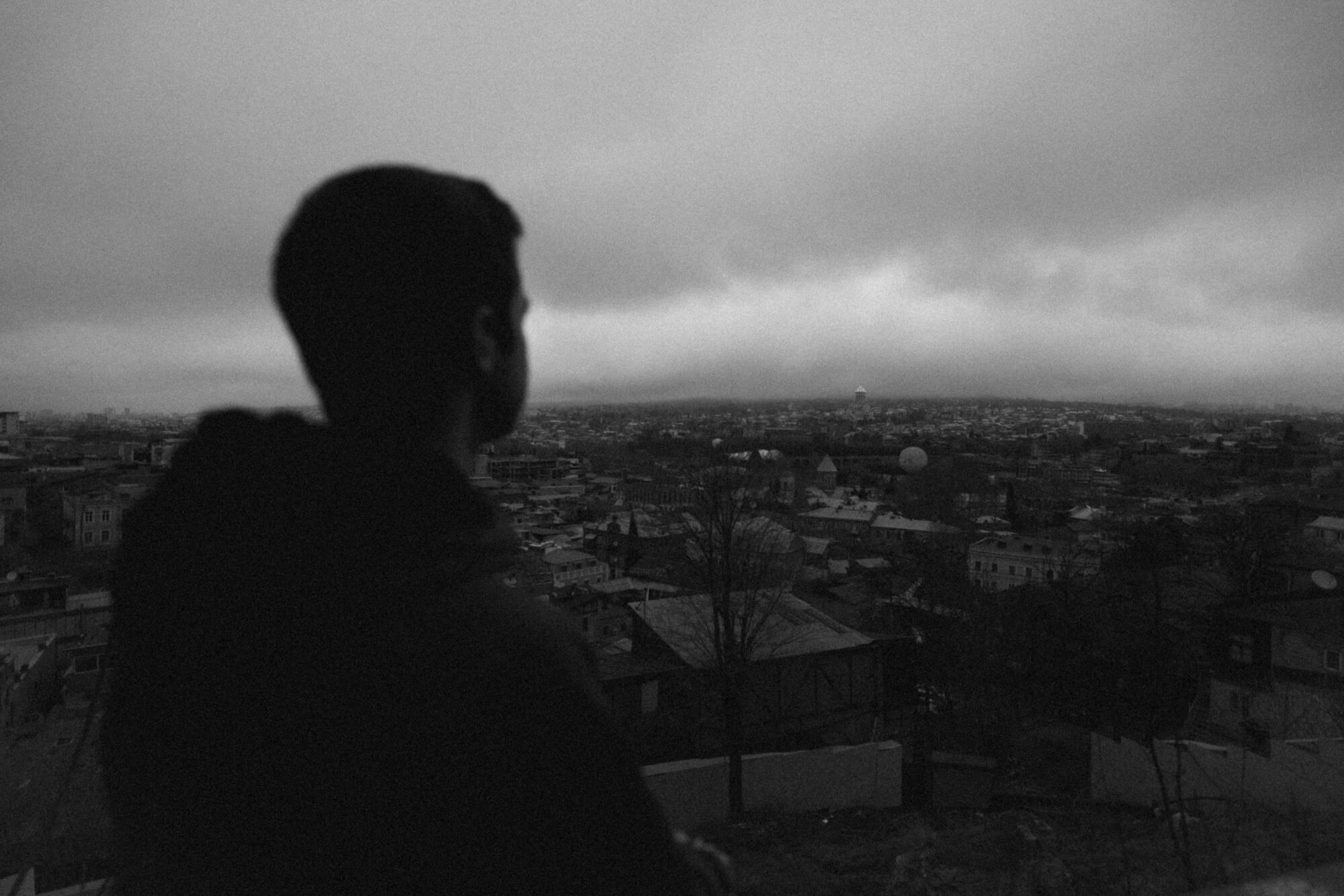 A person in silhouette looking out over a city skyline under a cloudy sky, with various buildings visible in the distance.