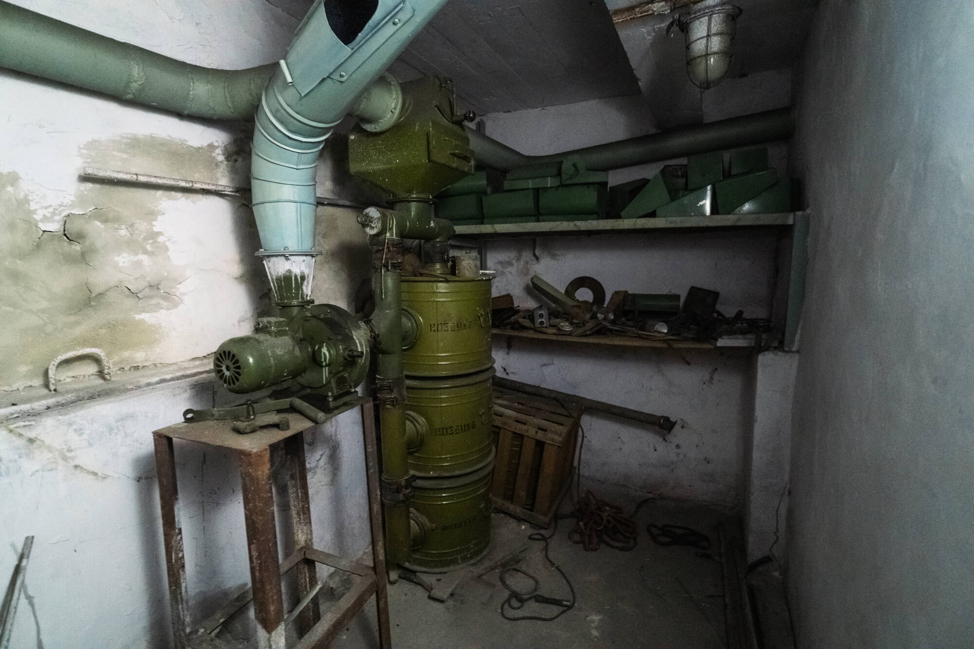 A cluttered industrial room featuring a green machine on a rusted stand, surrounded by pipes and various mechanical parts on shelves and the floor. The walls show signs of wear with cracks and peeling paint.