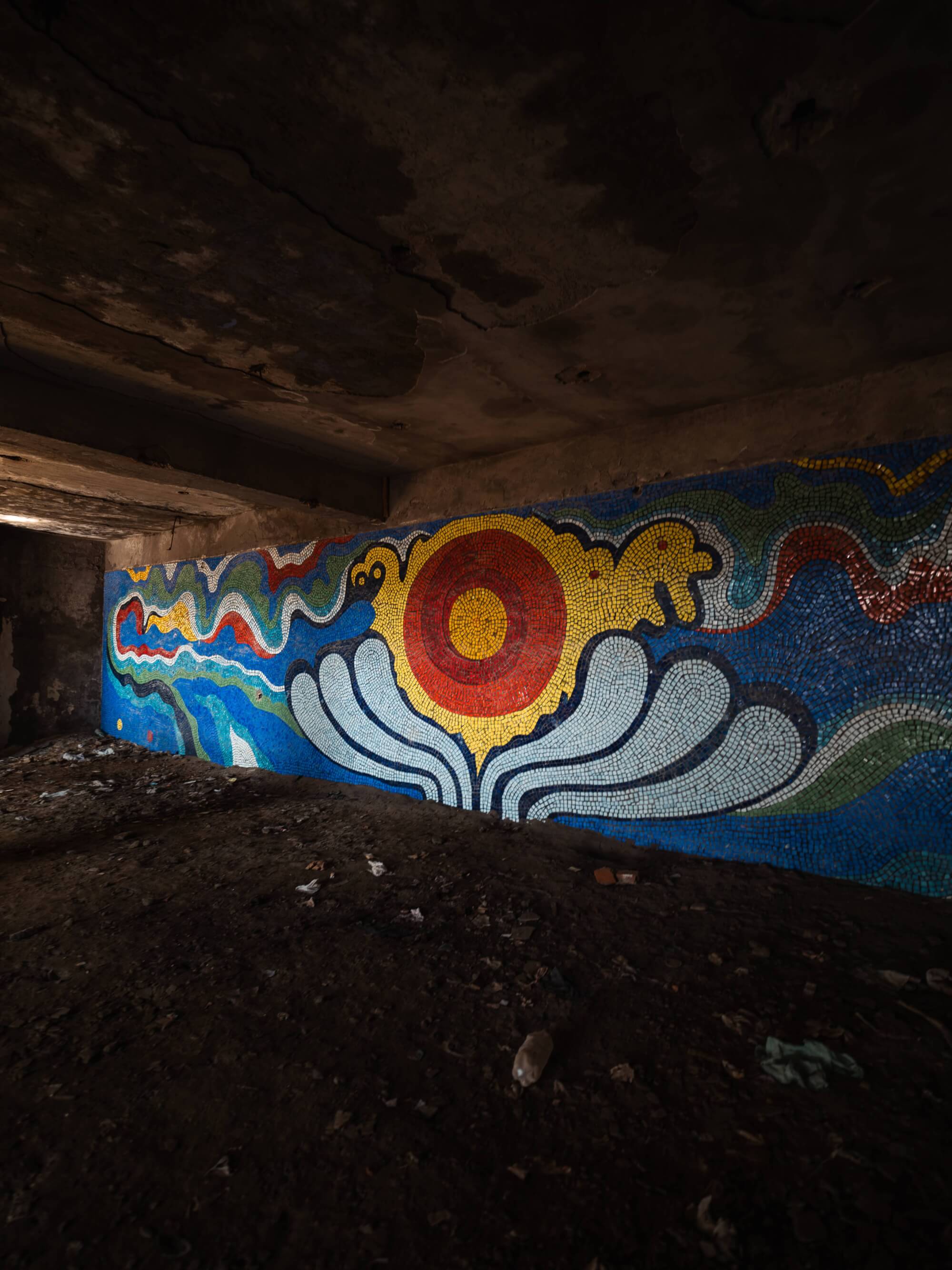 A colorful mosaic mural featuring a sun and flowing patterns, set against a dark, abandoned space with debris on the ground.