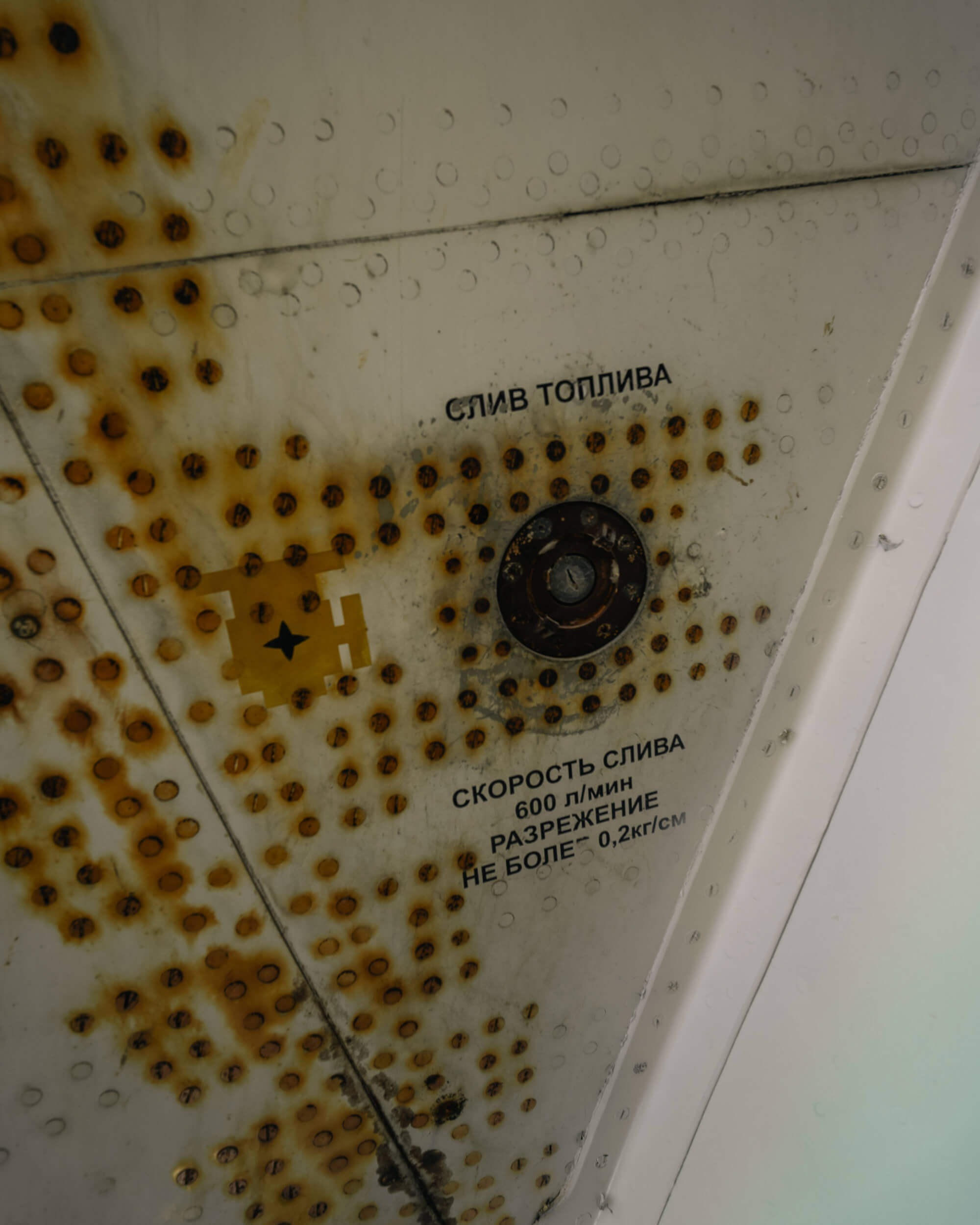 A close-up view of a rusted metallic panel with industrial markings, showing signs of wear, including brown rust spots. The panel contains inscriptions in Russian, indicating operational instructions, with a circular valve visible in the center.