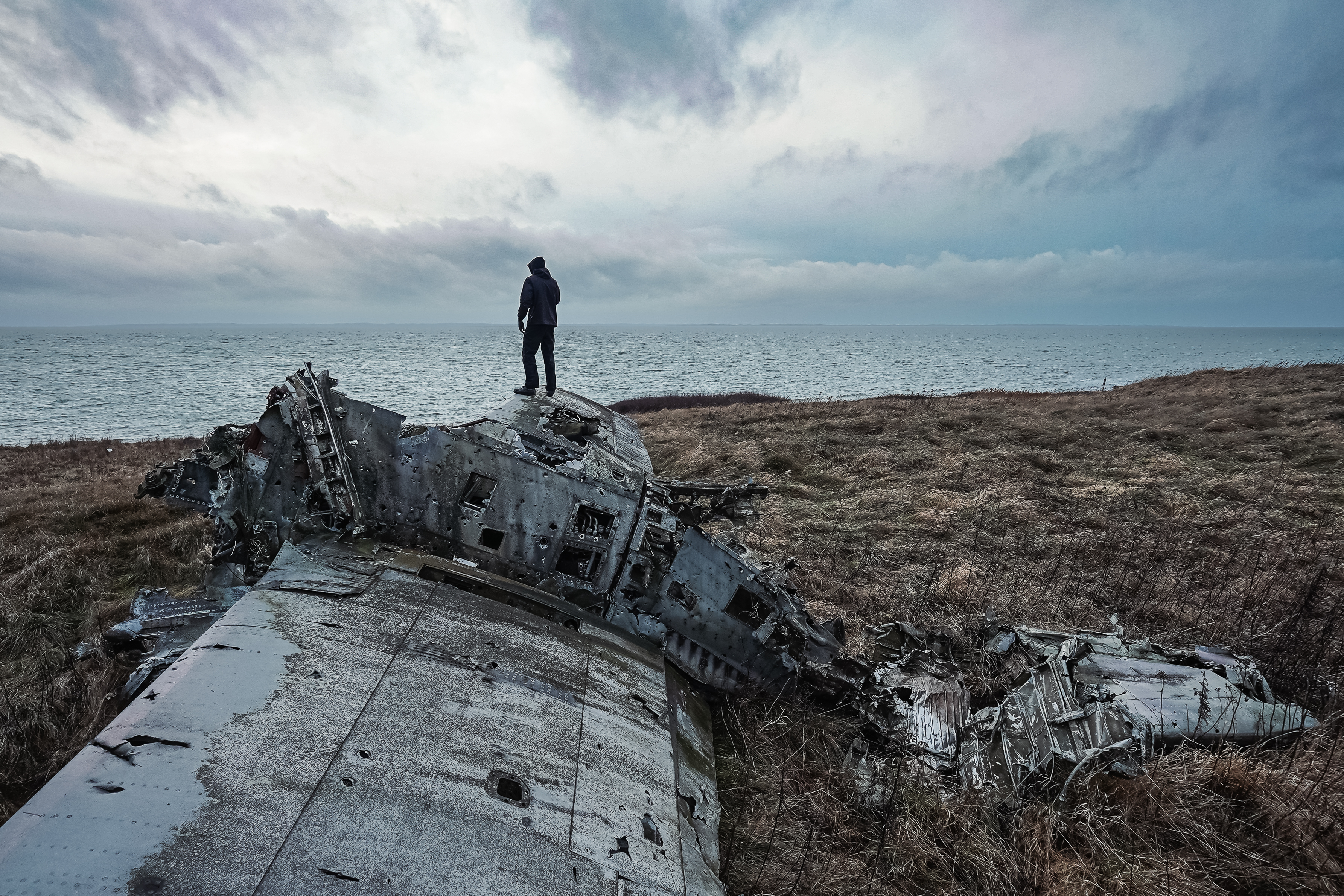 A person in a dark hoodie standing on the wreckage of a crashed airplane, overlooking the ocean under an overcast sky. The scene conveys a sense of solitude and abandonment.