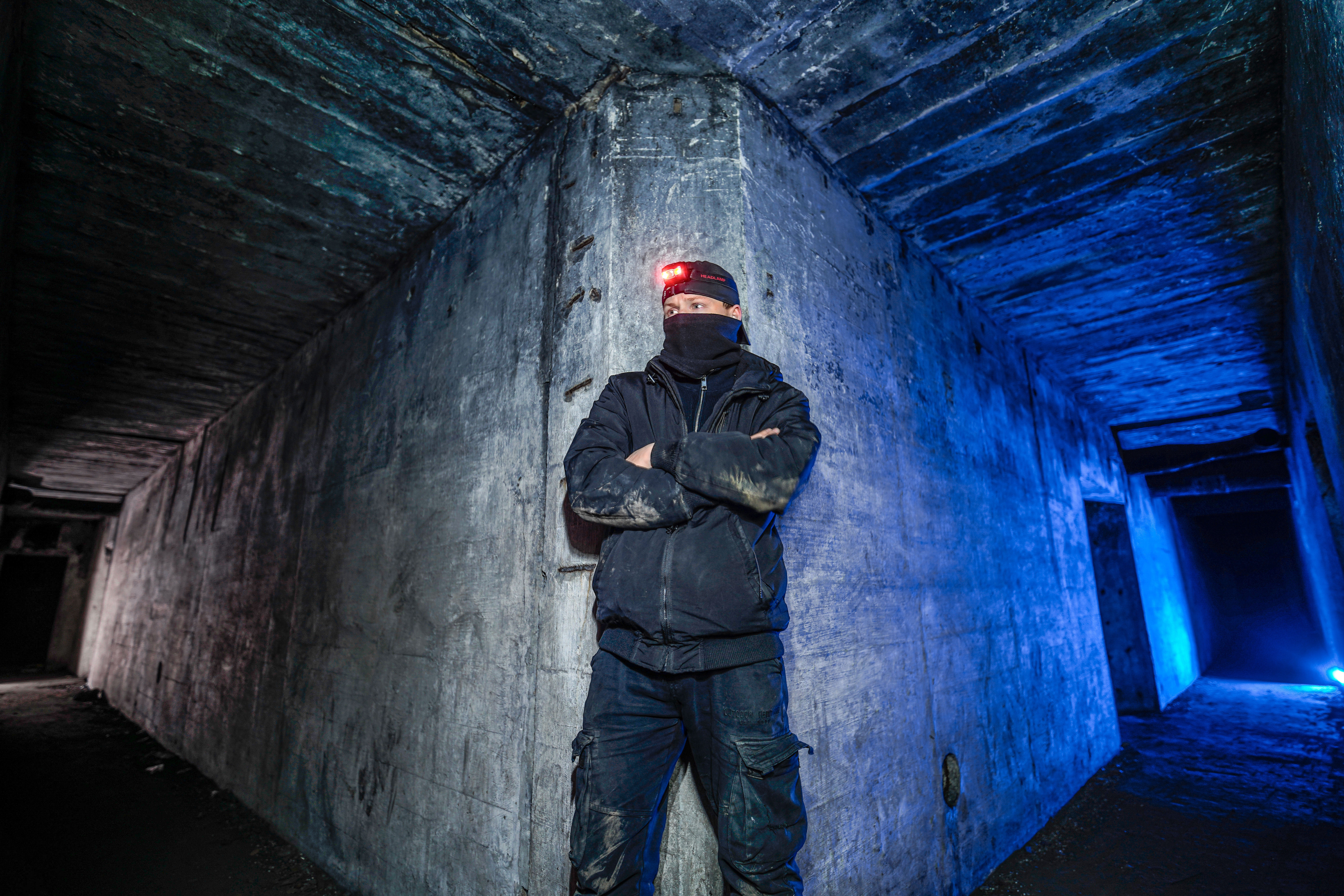 A person wearing a black jacket and pants, with a headlamp on their forehead, stands against a concrete wall in a dimly lit corridor. The area is illuminated with blue light, creating a dramatic contrast with the dark surroundings.