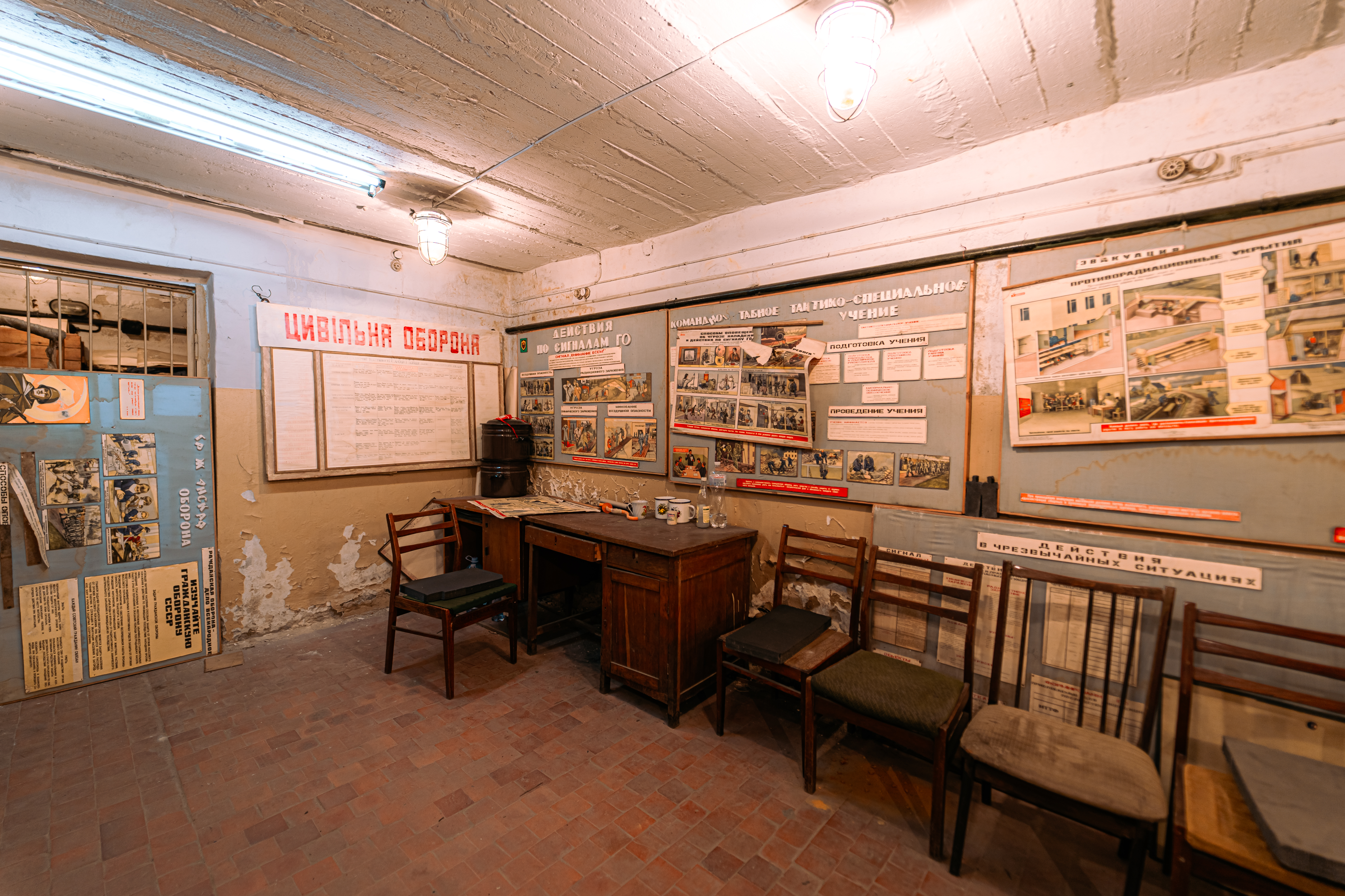 An interior view of a vintage civil defense training room with old wooden furniture, several walls covered in faded posters, and a desk cluttered with items. The lighting is soft and muted, creating a historical ambiance.