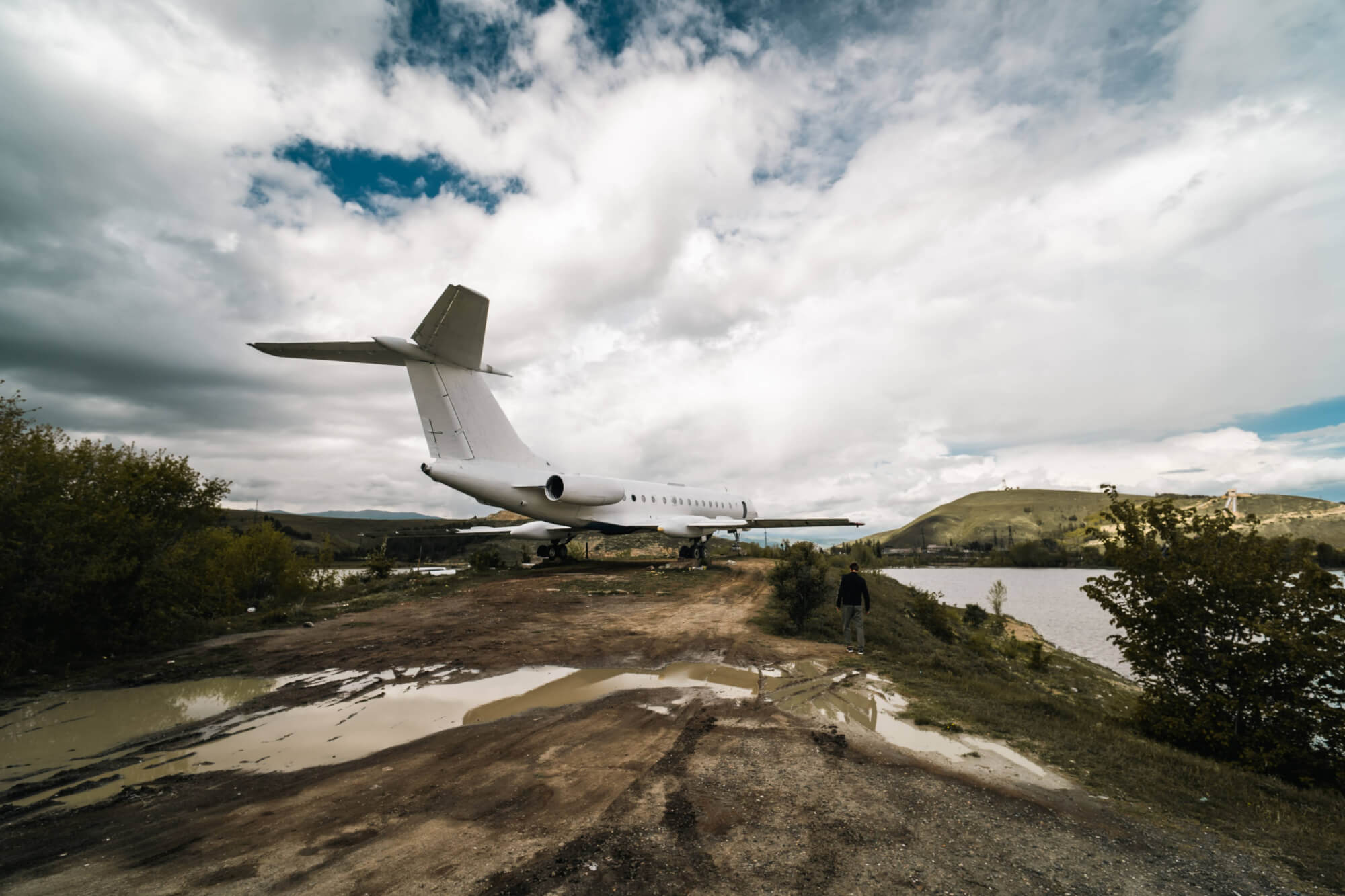 An abandoned white airplane on a muddy landscape with puddles, under a cloudy sky. A person walks beside a river in the foreground.