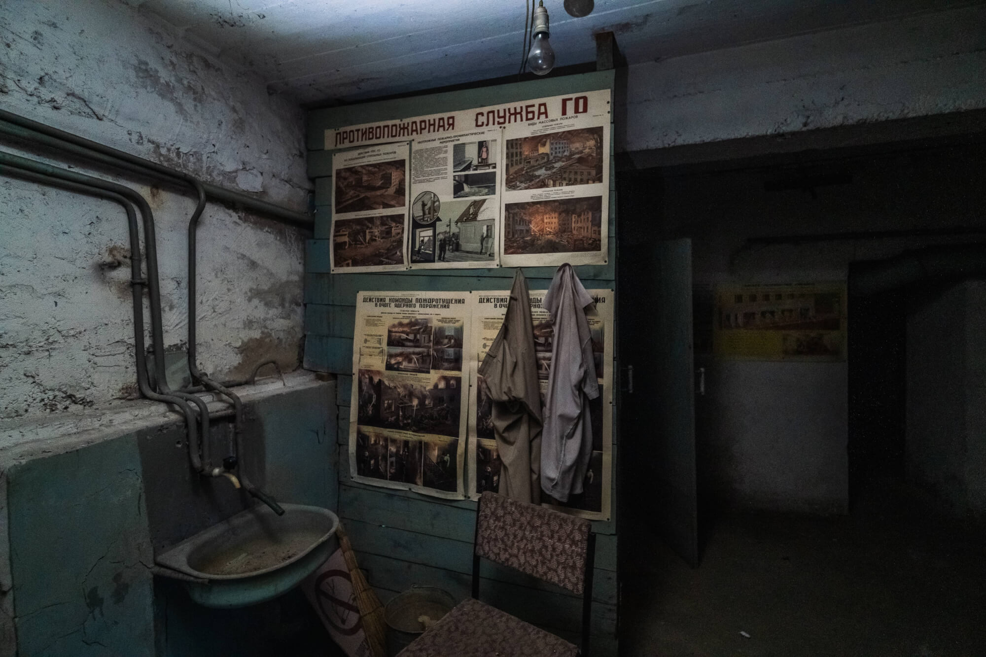 An empty, dimly lit room with a vintage fire safety poster on a peeling blue wall, a rusty sink, a chair, and two hanging uniforms.