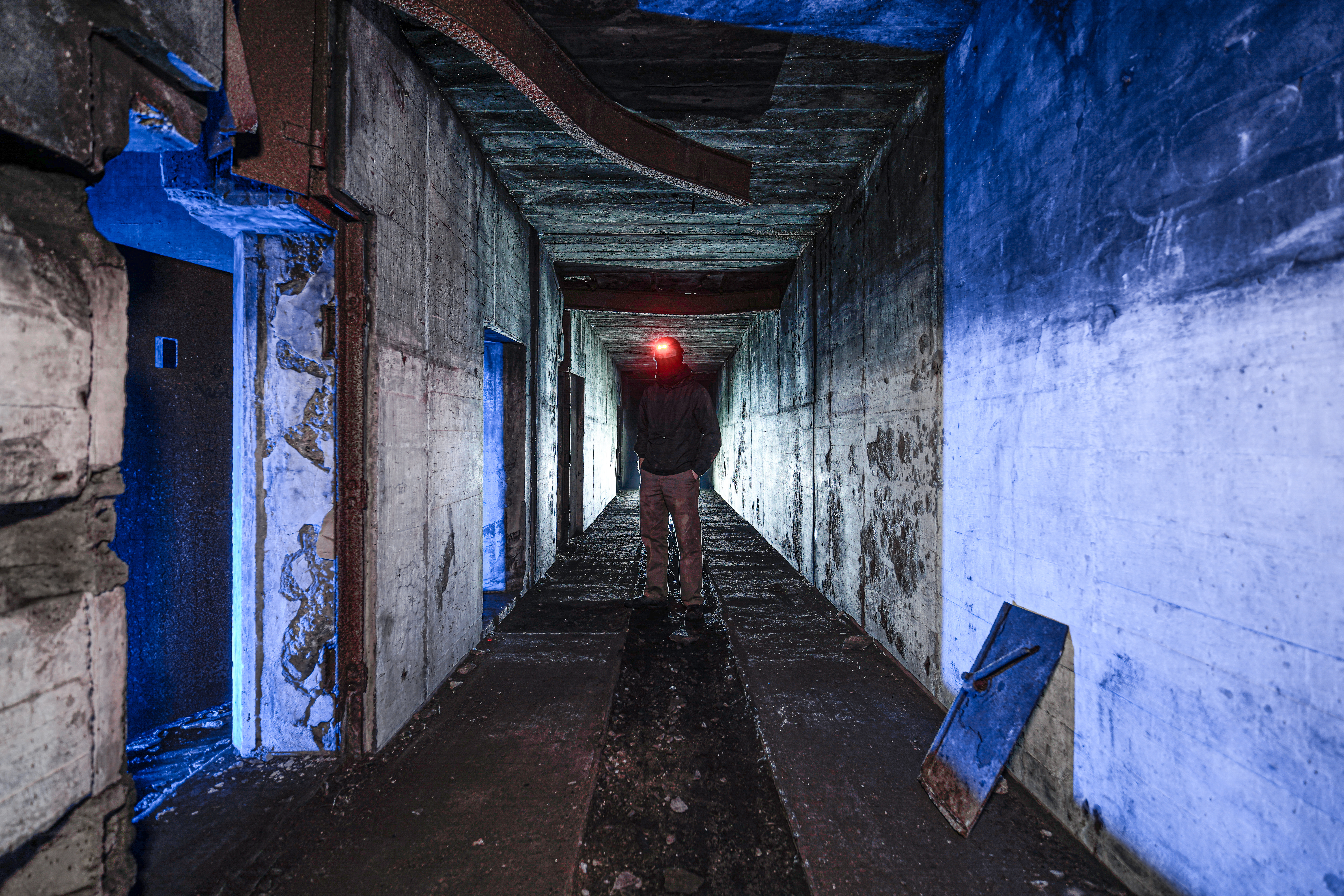A person in a dark hoodie stands at the end of an empty concrete hallway, illuminated by blue lights from side doorways and a red headlamp.
