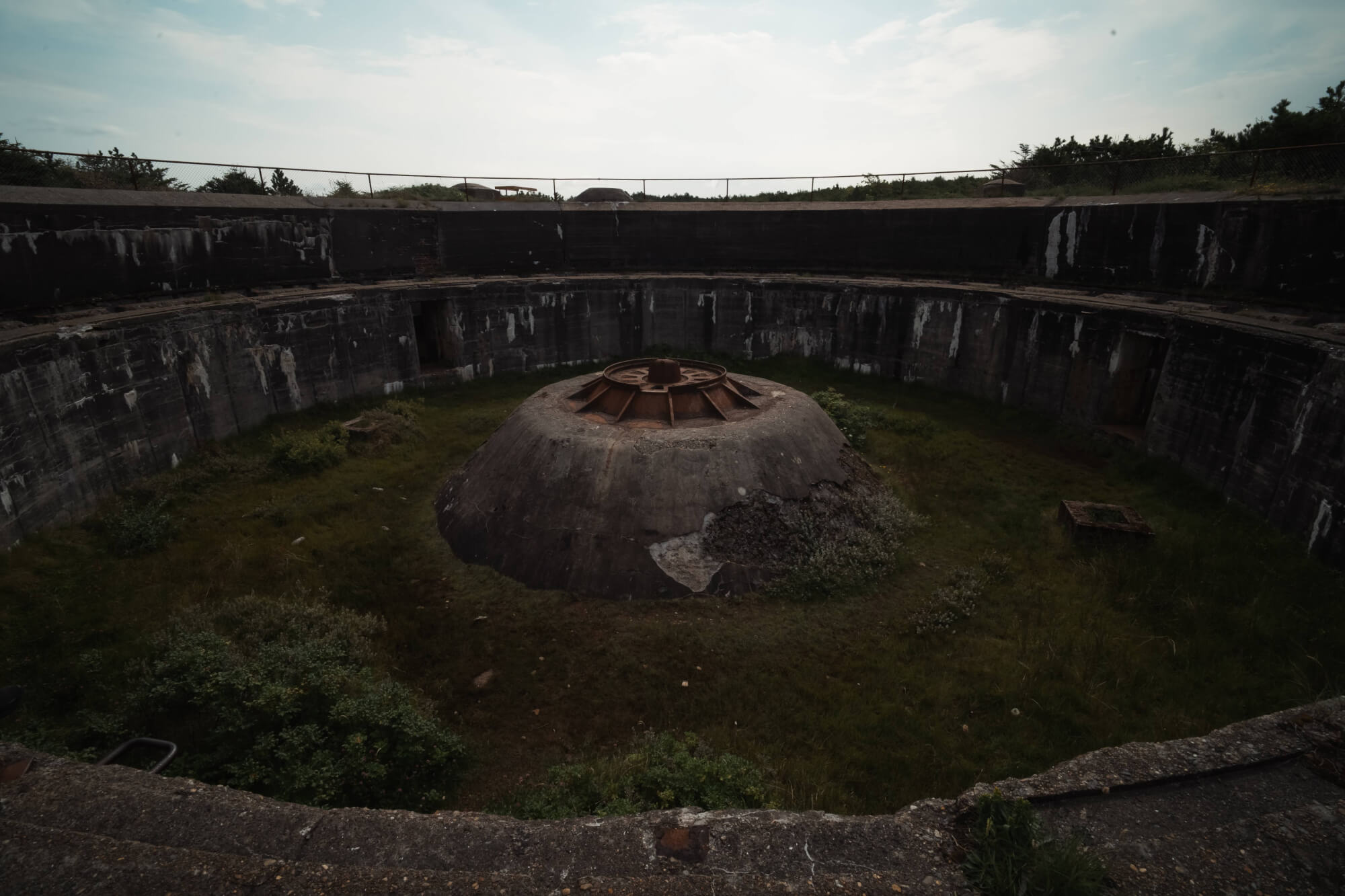 A large, dome-shaped concrete structure surrounded by overgrown grass, situated in a circular concrete chamber with weathered walls. The sky is cloudy, adding a moody feel to the scene.