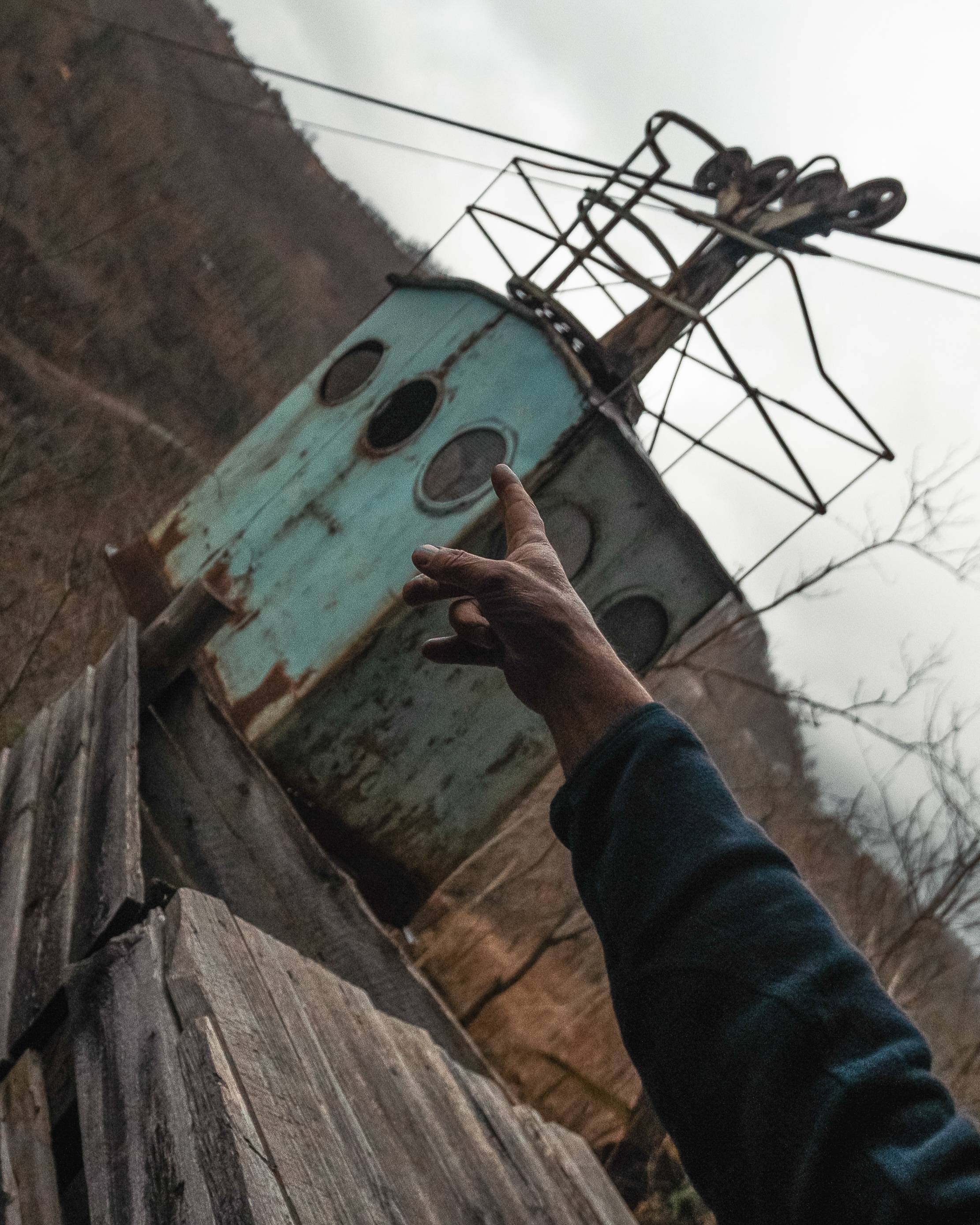 A weathered blue cable car on a cable line with a hand pointing towards it. The background features bare trees and a cloudy sky, creating a somber atmosphere.