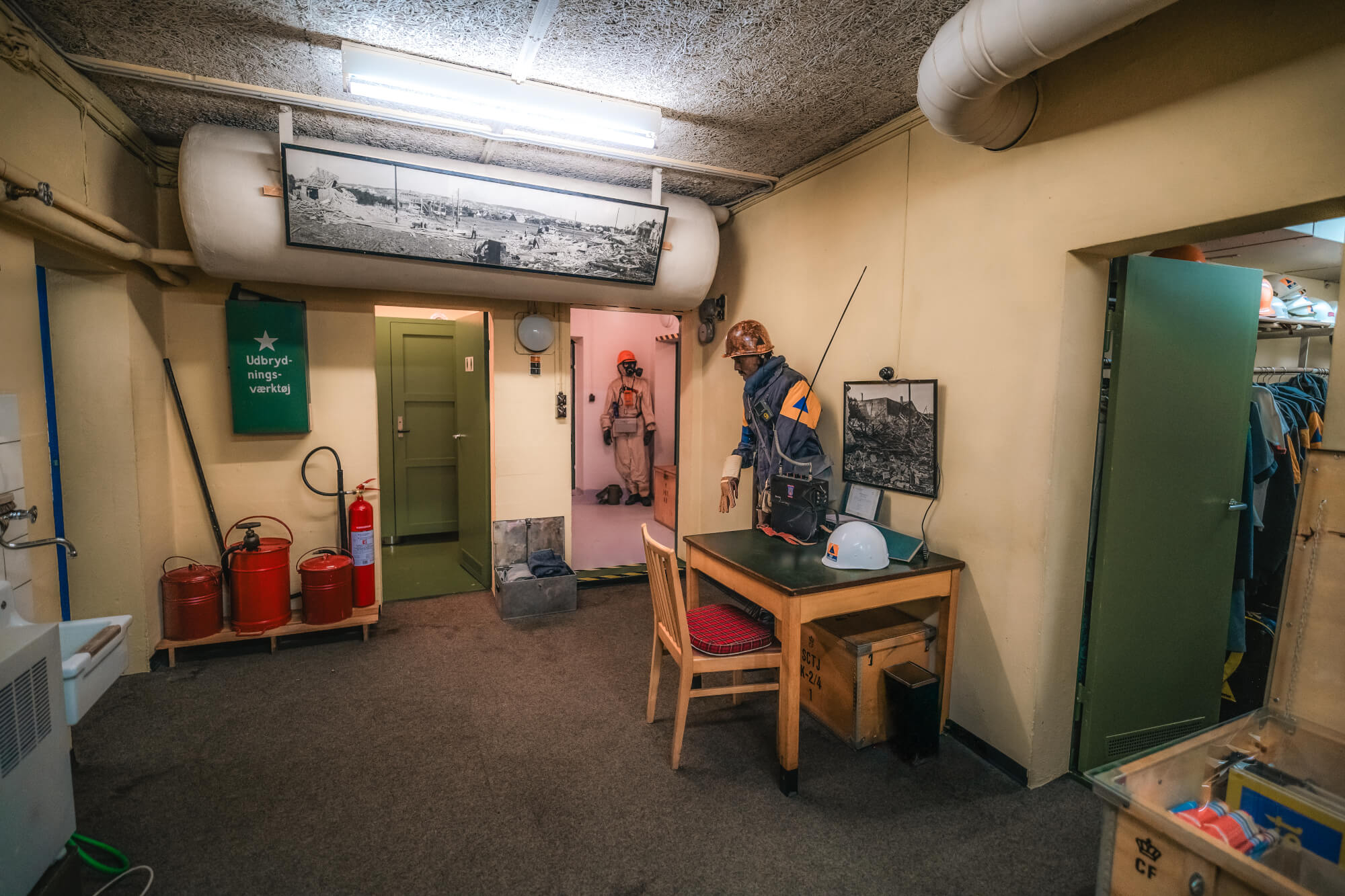 An underground room featuring a wooden table and a mannequin in work attire. A doorway leads to a figure in a light-colored suit, with fire extinguishers and various utility items positioned around the room.