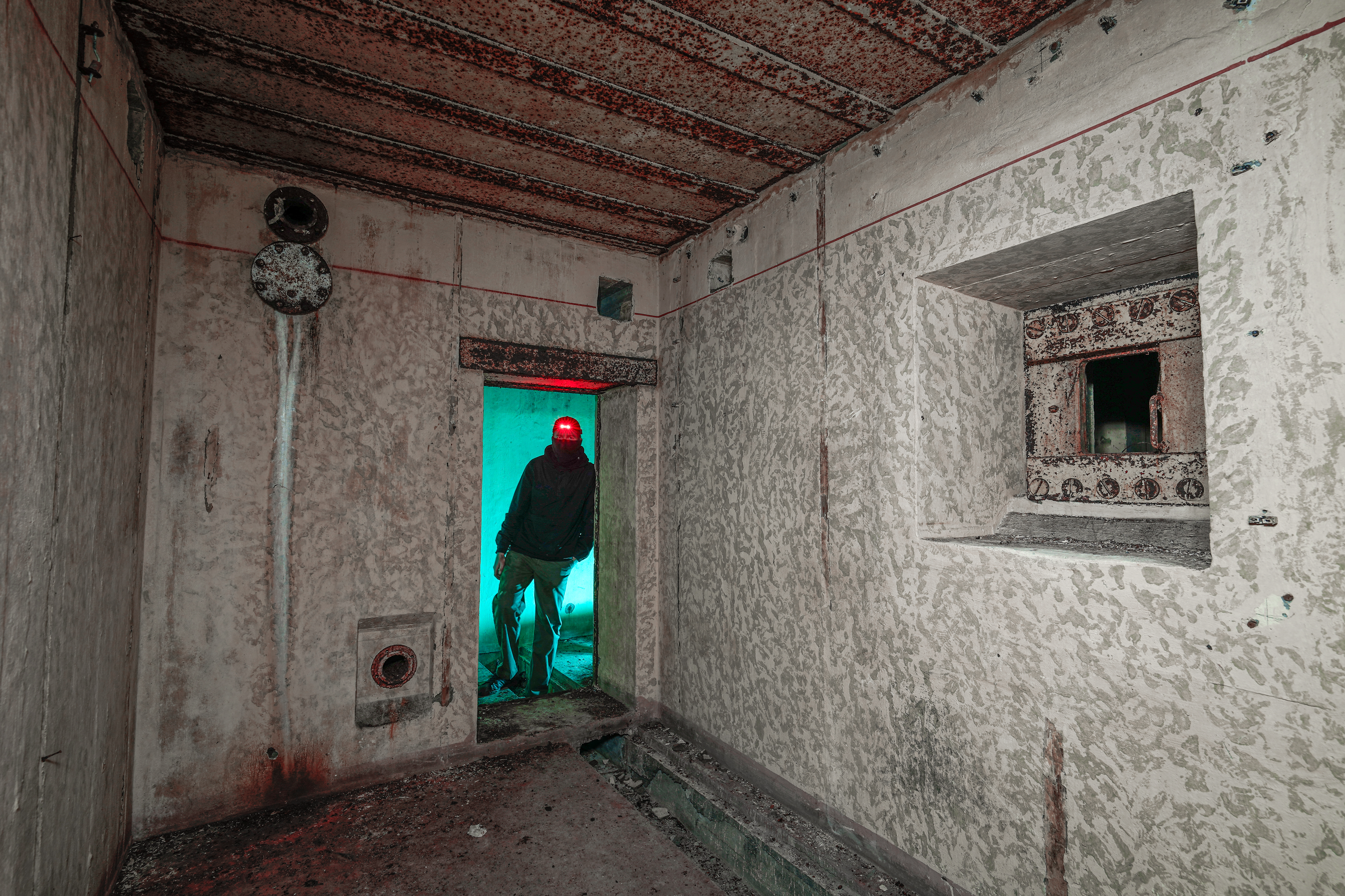 An abandoned room with peeling walls and rust, featuring a person in dark clothing standing in a doorway with blue and red lighting.