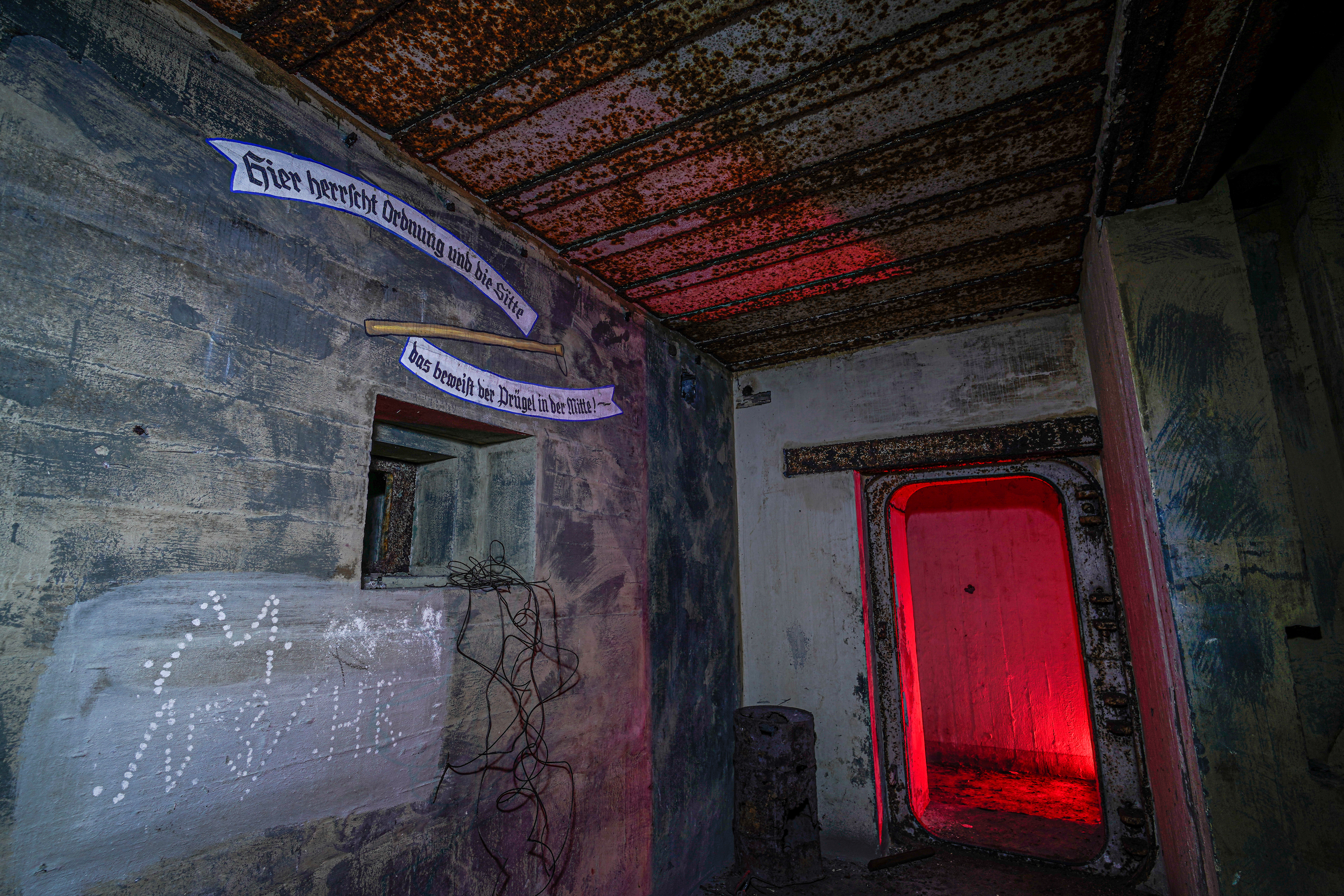 An abandoned room with rusty ceilings, peeling paint, and tattered banners on the walls. A doorway with a red glow leads to another space, while twisted wires and faint graffiti contribute to the eerie atmosphere.