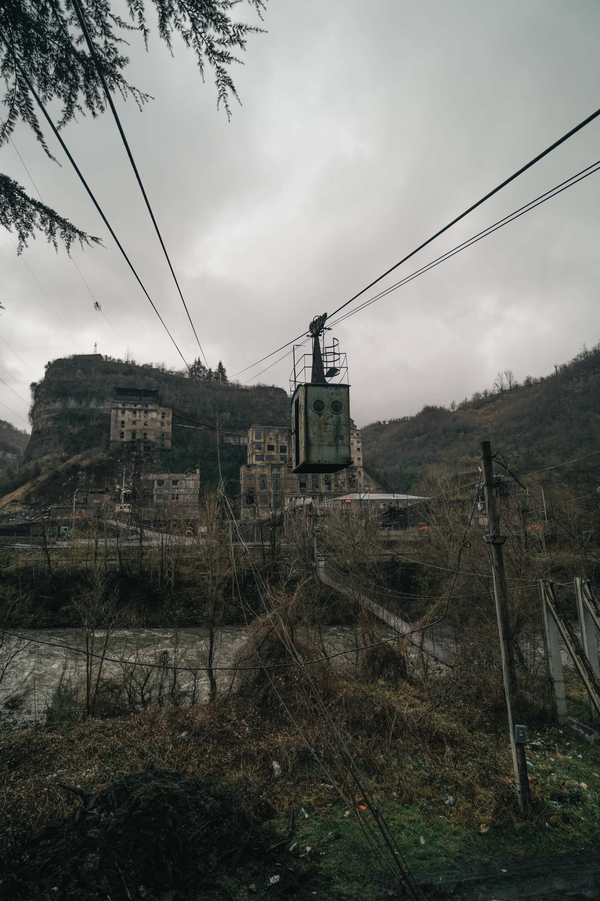 An old cable car hangs over a river, with abandoned buildings and a rugged hill in the background under a cloudy sky.