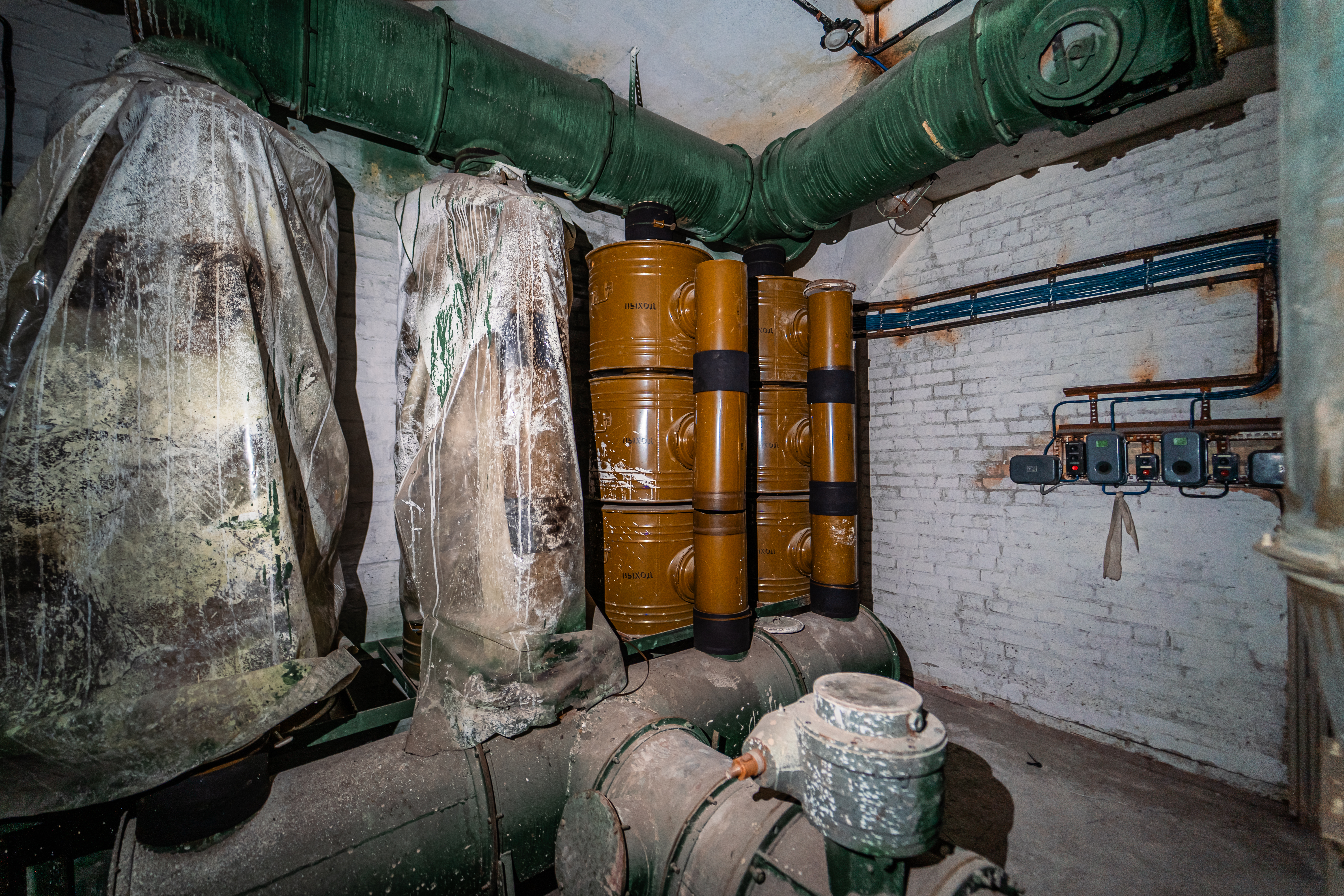An industrial room with large green pipes, two covered structures wrapped in plastic, and several yellow barrels stacked against a white brick wall. Blue electrical wires and multiple meters are visible on the wall.
