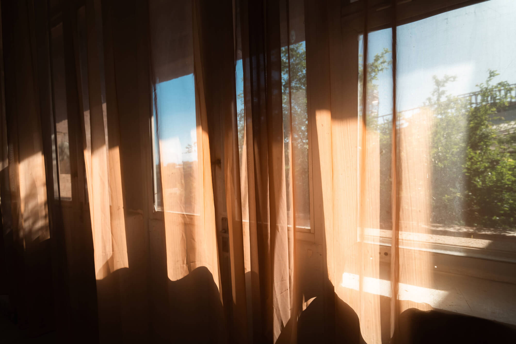 A room interior with sheer curtains partially draping a window. Sunlight streams through, illuminating the fabric and casting soft shadows. Outside, greenery is visible, suggesting a tranquil natural setting.