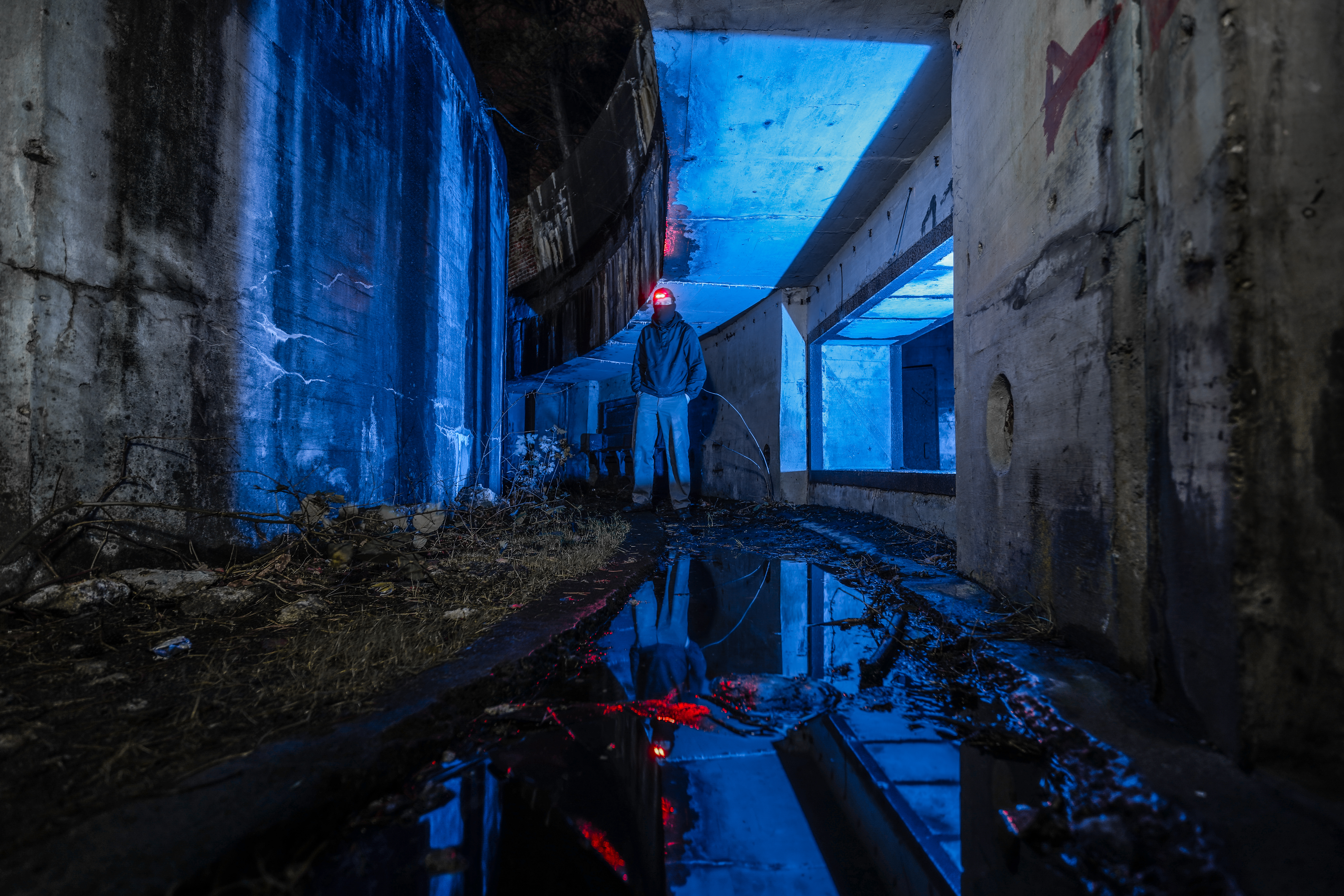 A person wearing a dark hoodie stands in an underground space with blue and red lighting, reflecting in a puddle on the ground. Concrete walls create a stark environment, with sparse vegetation visible.