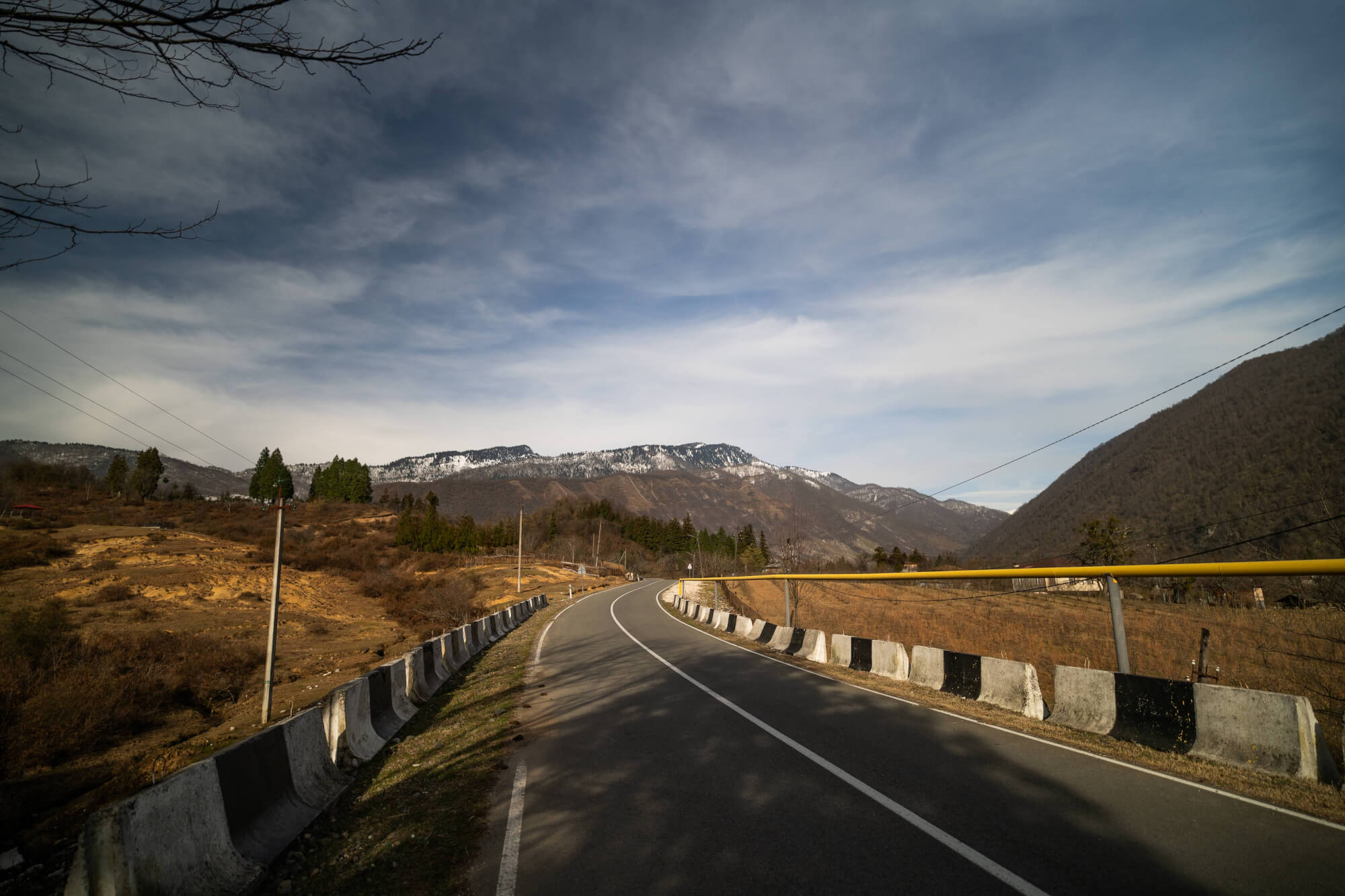 A winding road with black and white barriers alongside, leading toward distant snow-capped mountains under a partly cloudy sky.