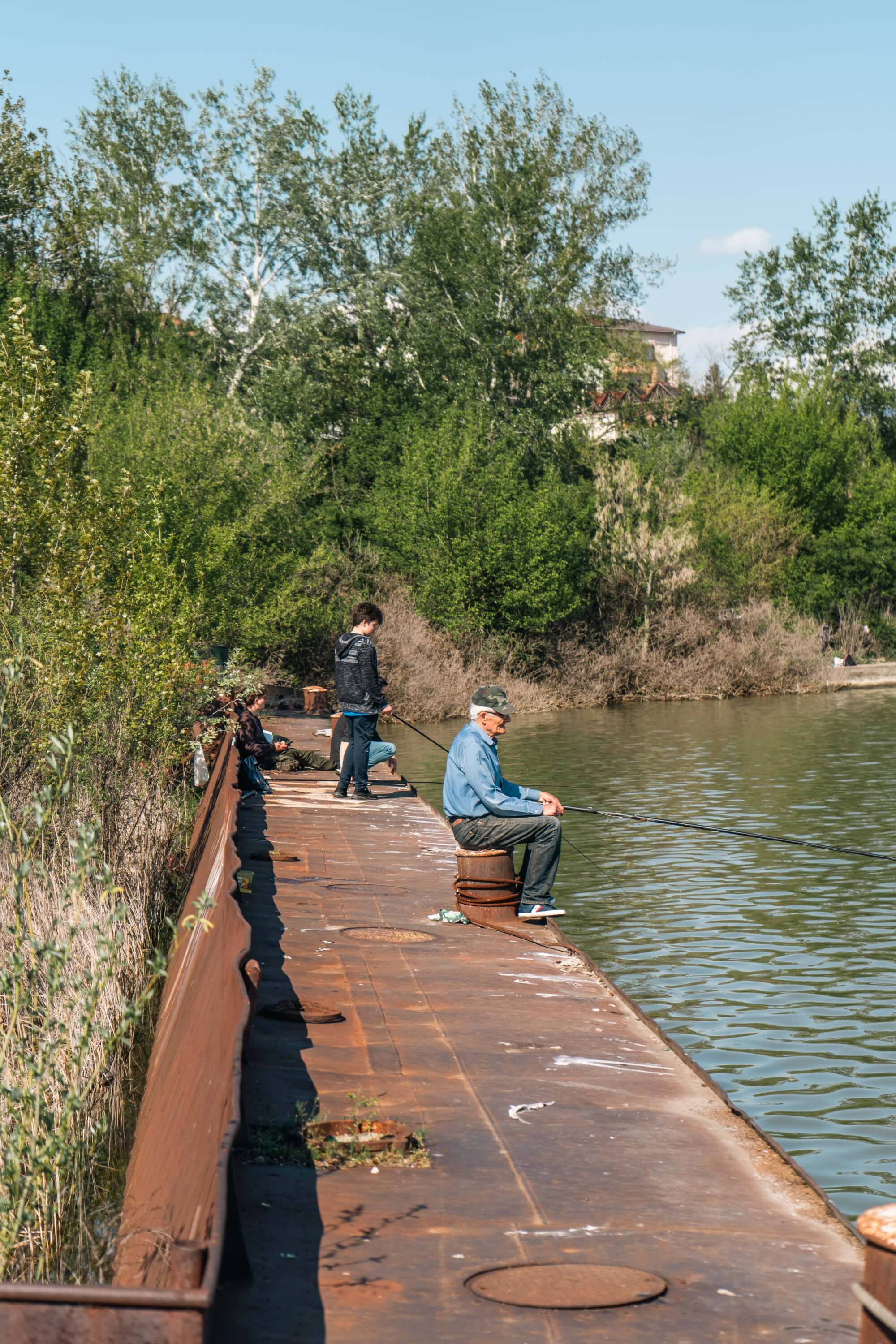 Several people fishing on a rusty pier by a calm body of water, surrounded by green trees and shrubs.