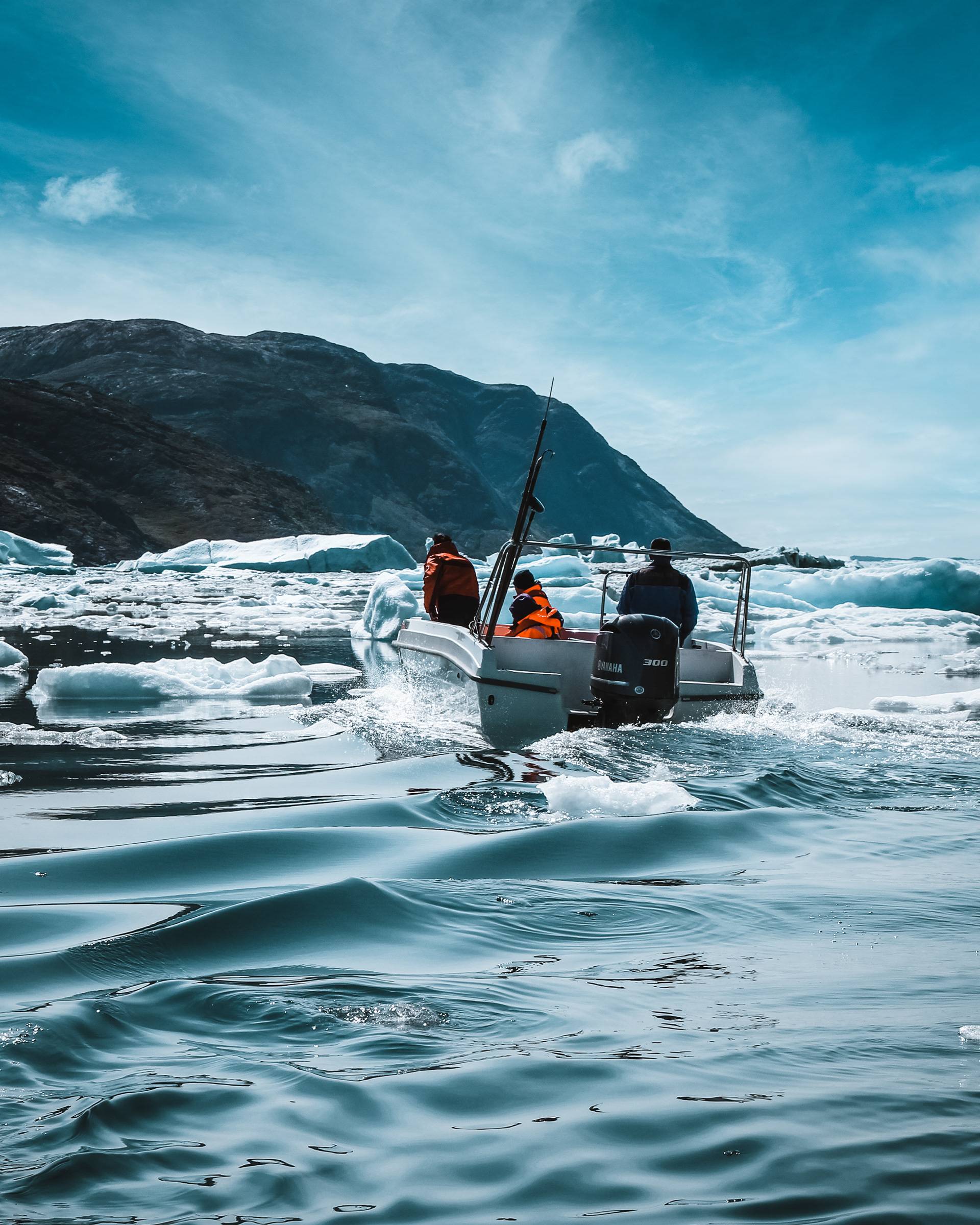 A small boat with three passengers wearing bright orange life jackets is navigating through icy waters, with scattered icebergs and a mountainous backdrop under a bright blue sky.