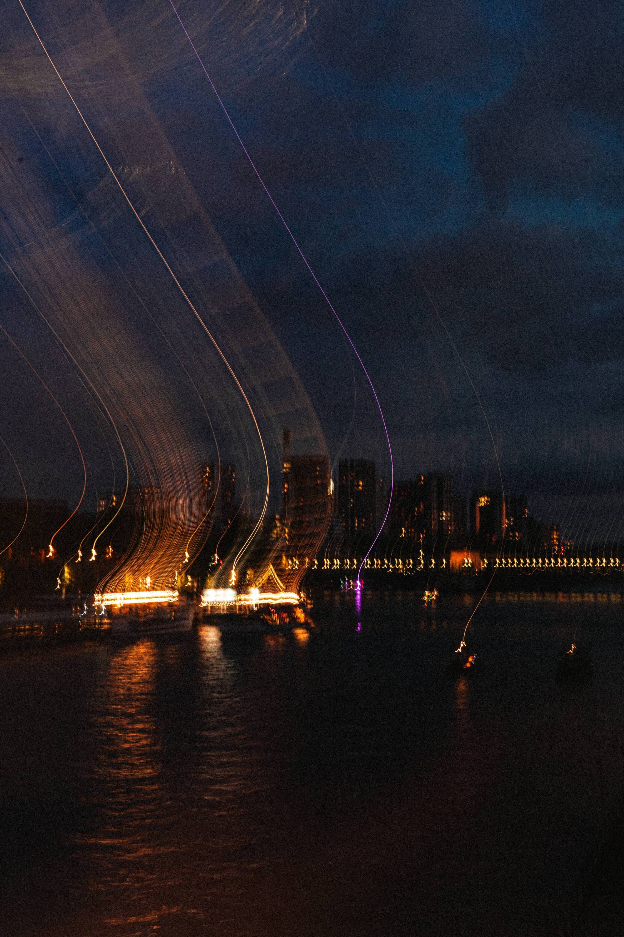 A long exposure photograph of a river at night with streaks of light from boats, high-rise buildings in the background, and a darkening sky.
