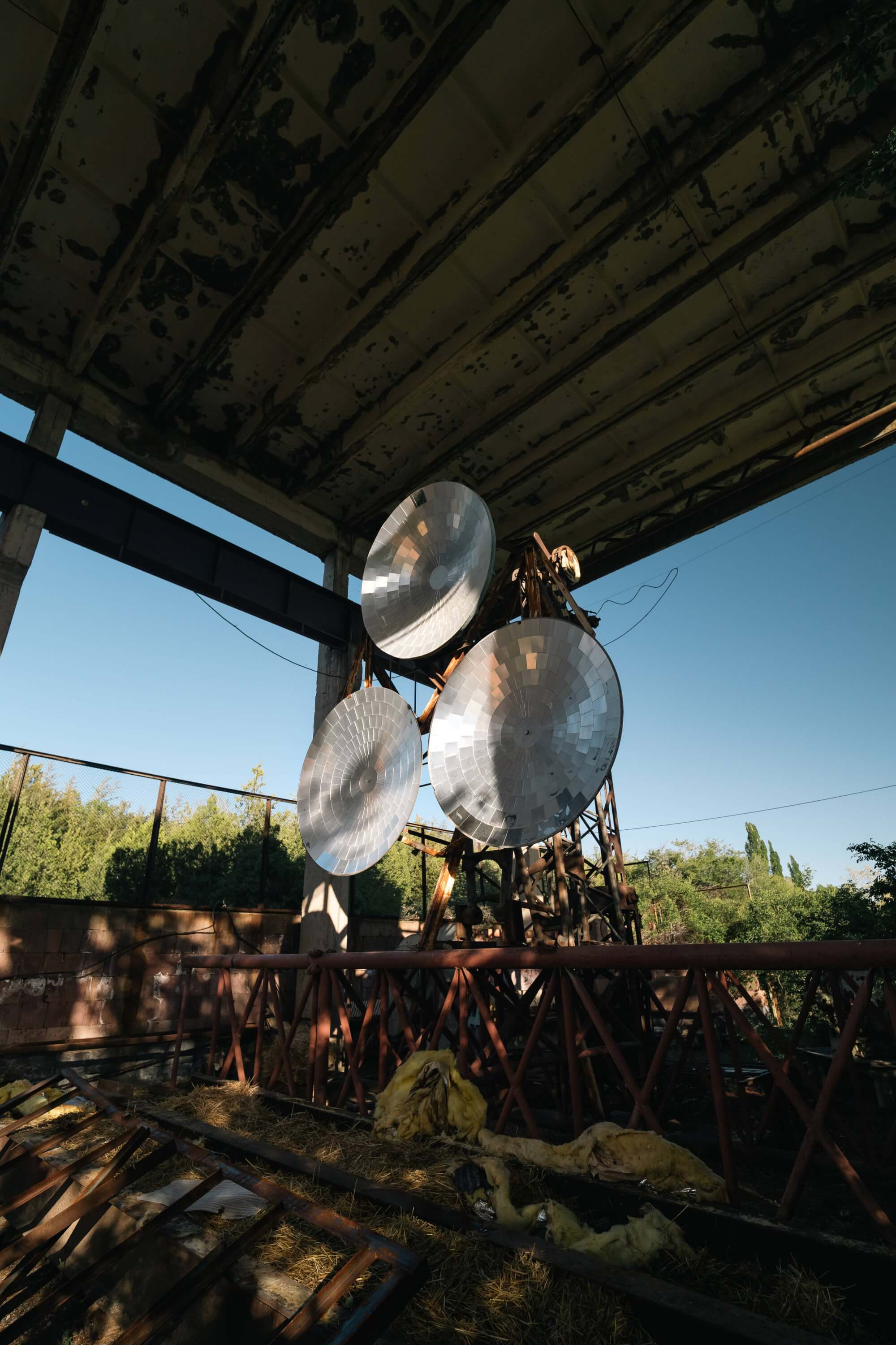 A view of three large metal dishes mounted on a rusted frame inside an abandoned building, with scattered debris and overgrown foliage visible through an open window.