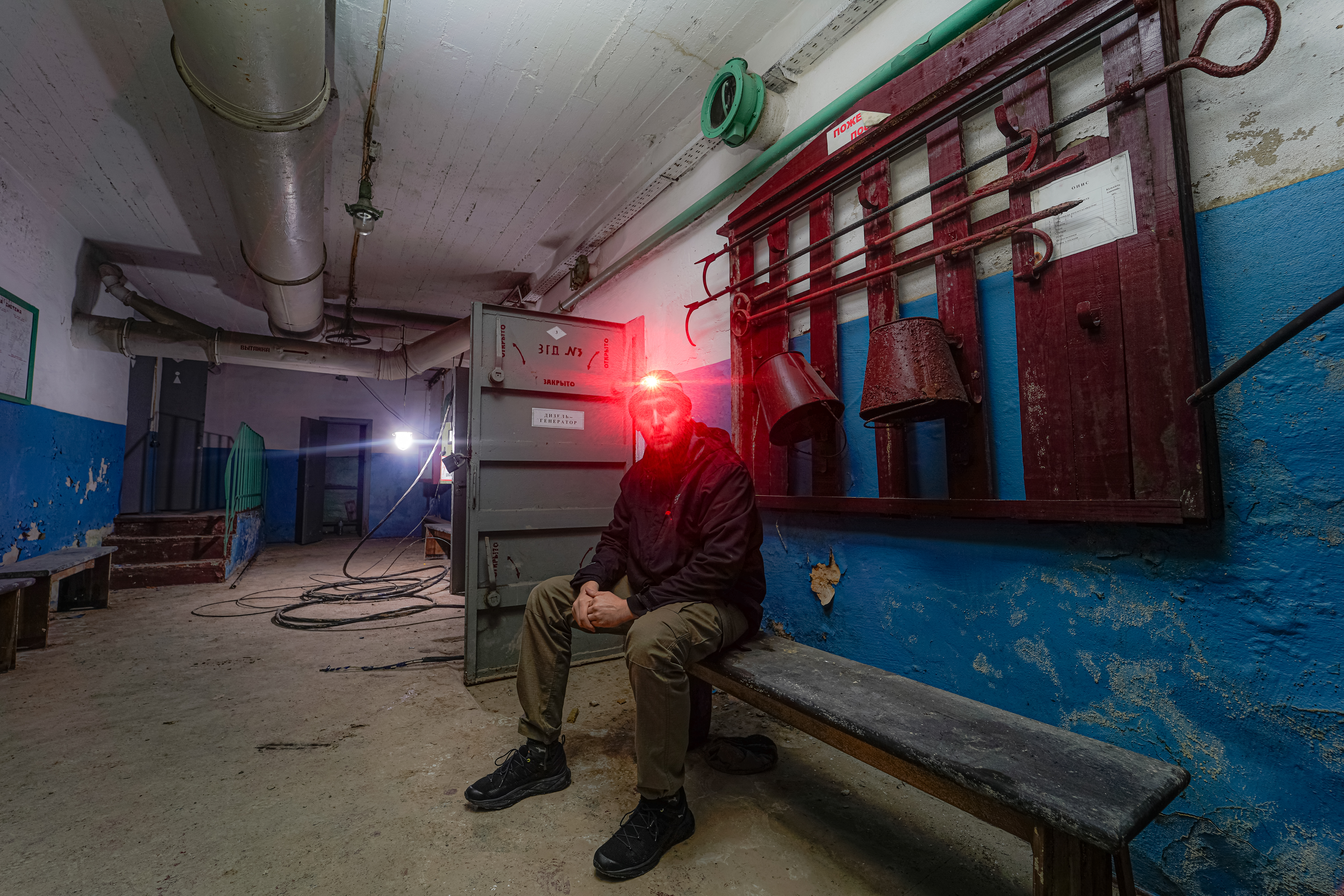 A man sitting on a bench in an abandoned hallway, illuminated by a red headlamp. The walls are blue and white, with a rusty red panel holding various tools. Background includes dim lighting and scattered debris.