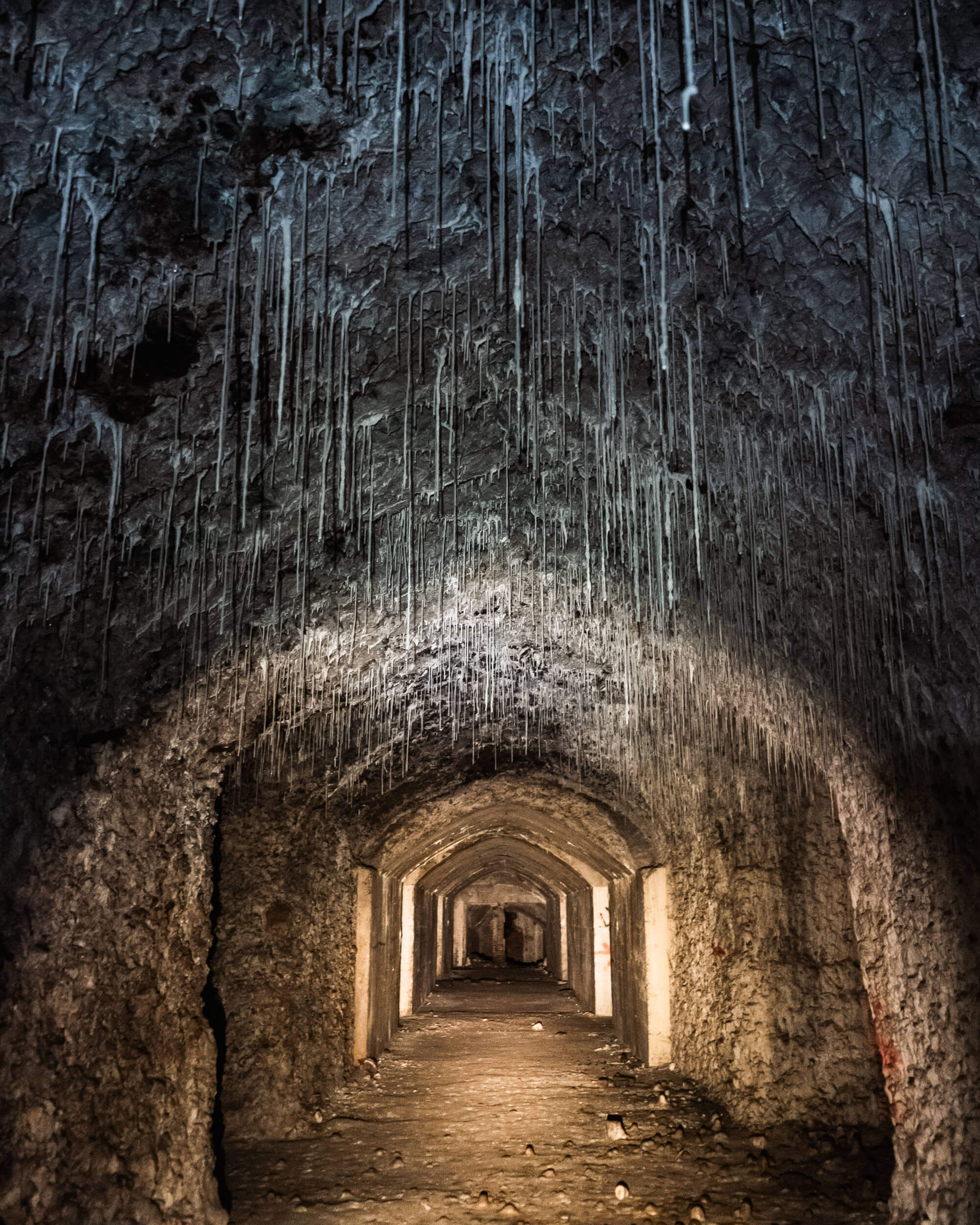 A dimly lit underground tunnel with rocky walls and a ceiling featuring long, thin icicle formations. The floor is uneven with scattered stones and debris, leading deeper into the tunnel.