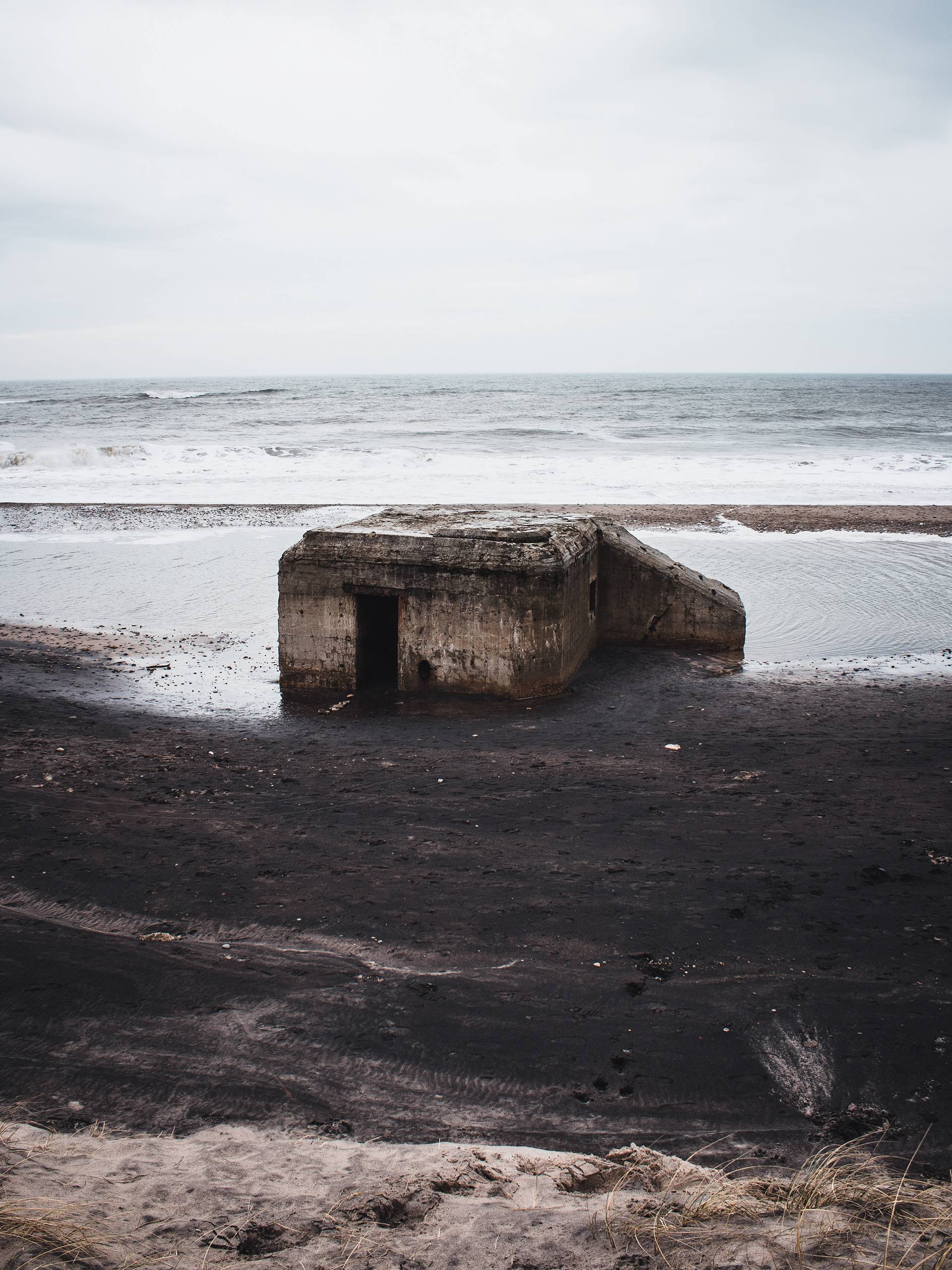 A concrete bunker on a dark beach near the ocean, with overcast skies. Waves are crashing in the background, and the sand is mostly dark with some light-colored patches.