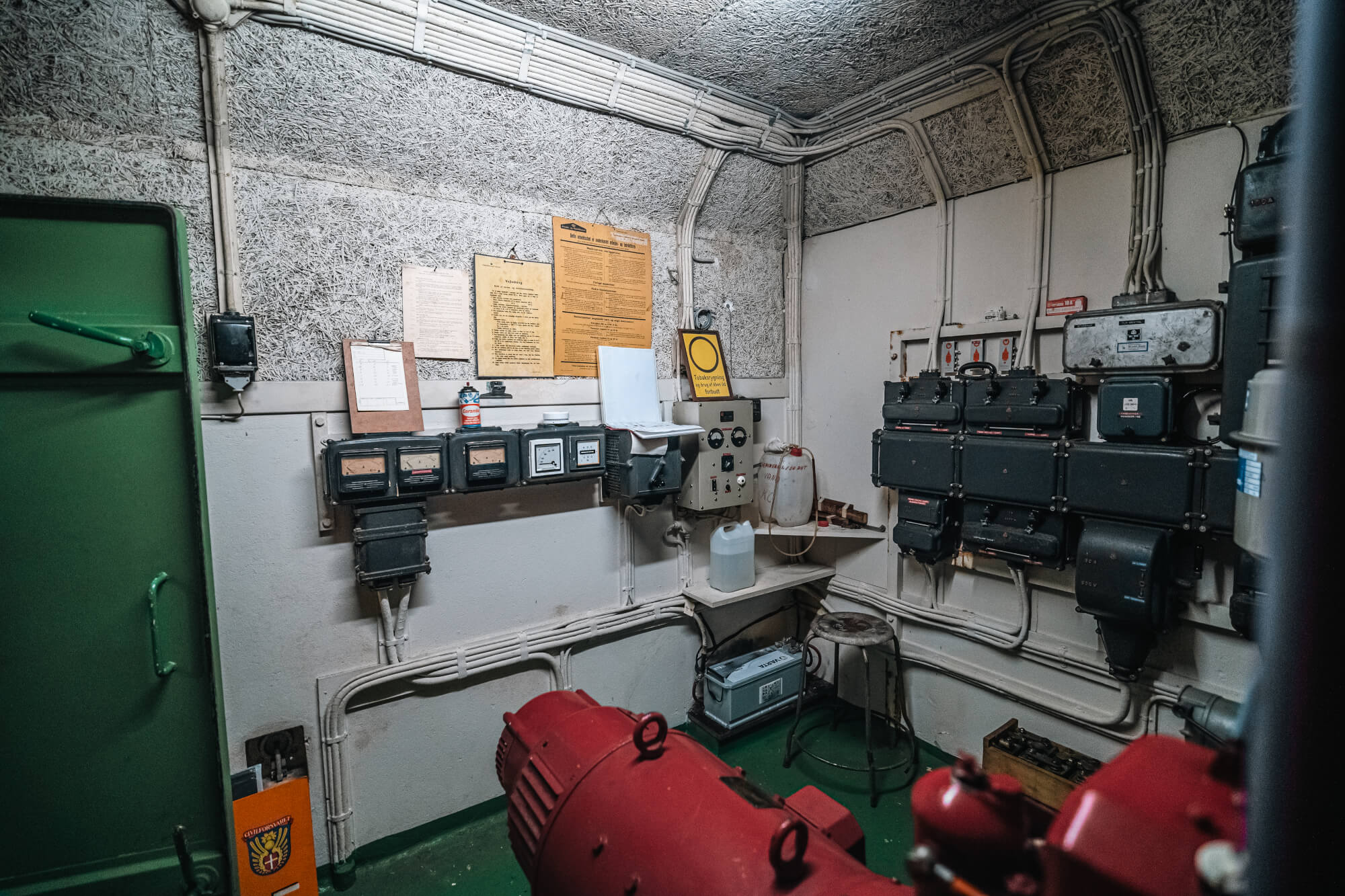 Interior of an industrial control room featuring a red device in the foreground, various meters and panels on the wall, and a stool beside a shelf. The walls are textured and cables are visible.