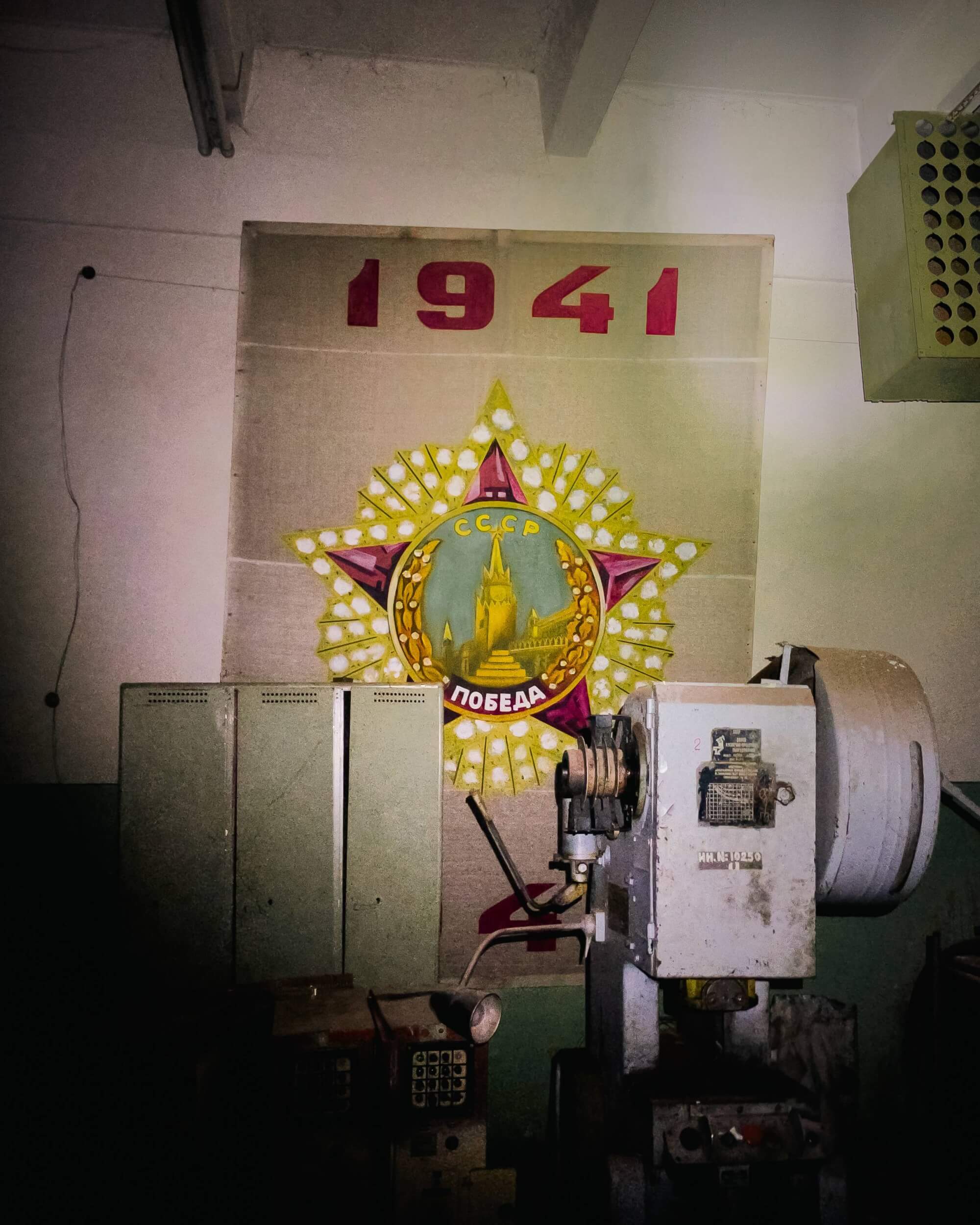 A vintage Soviet banner with the year '1941' is displayed on a concrete wall in a dimly lit industrial setting, accompanied by old machinery and green metal cabinets.
