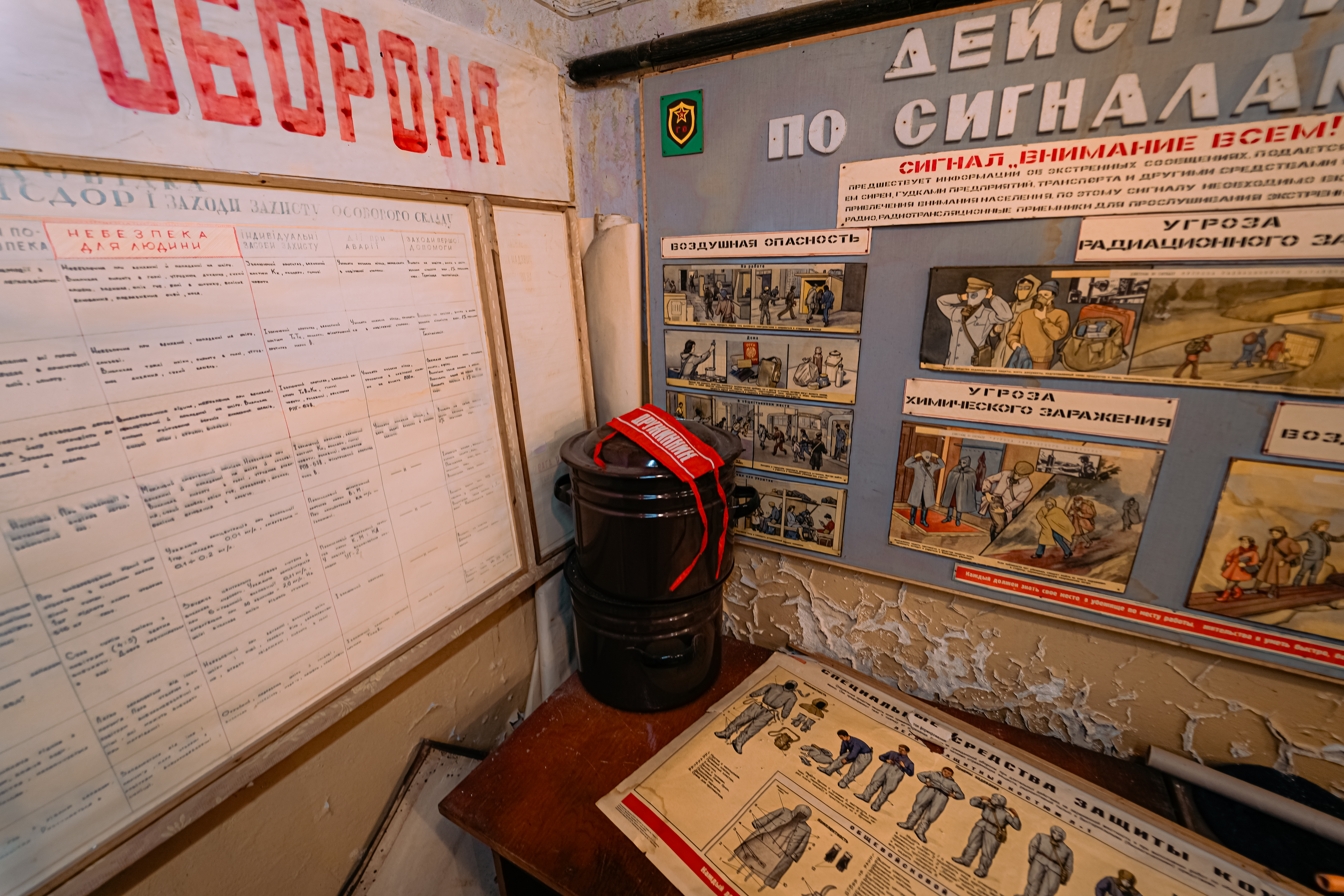An interior corner featuring a bulletin board with safety posters in various colors and a wooden table with two black containers and a red strap. The walls are faded and worn, showing signs of age.