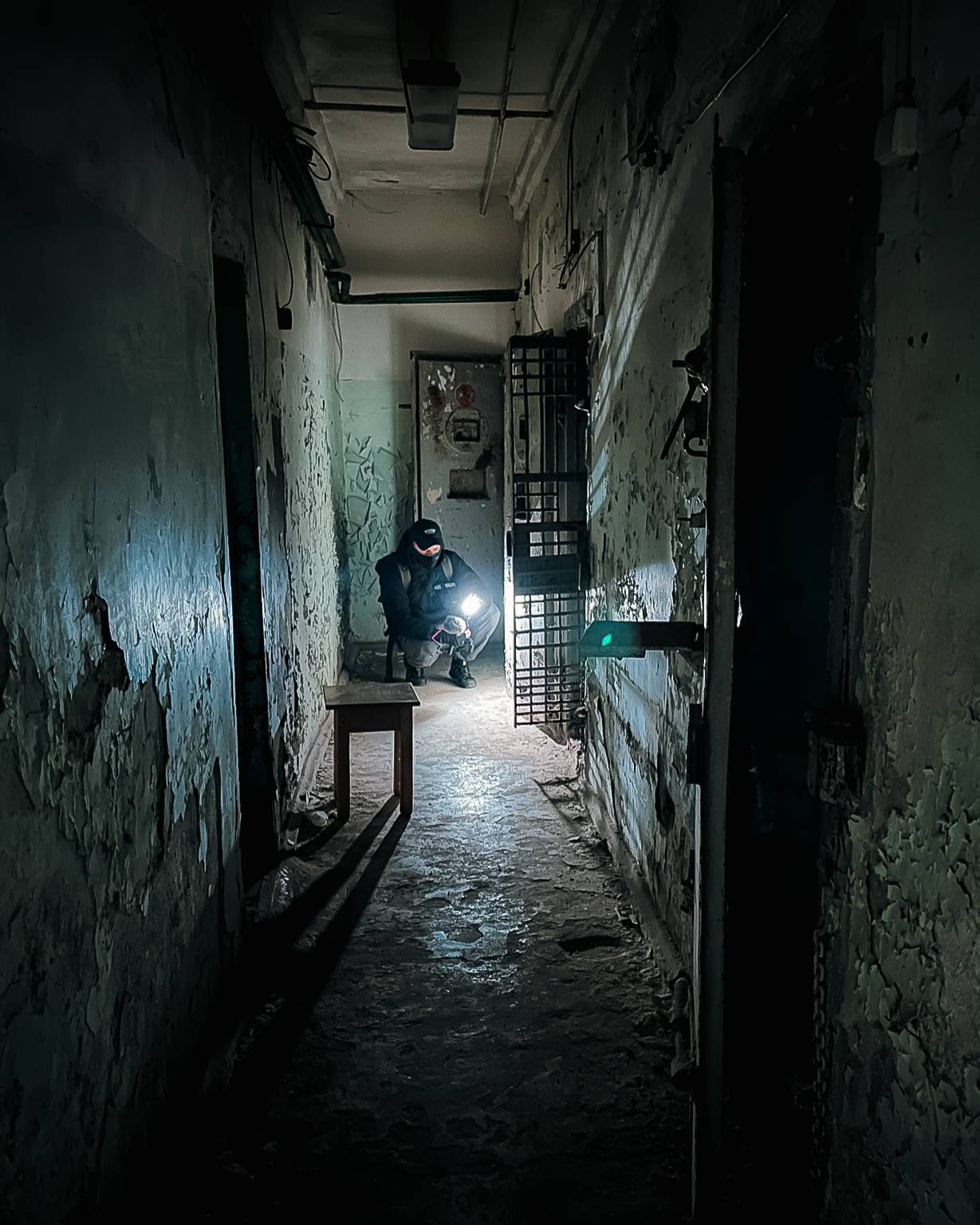 A dimly lit corridor in an abandoned building, featuring a figure crouched down holding a flashlight. The walls are peeling and distressed, with a table on the left casting a long shadow. A barred door is visible in the background.