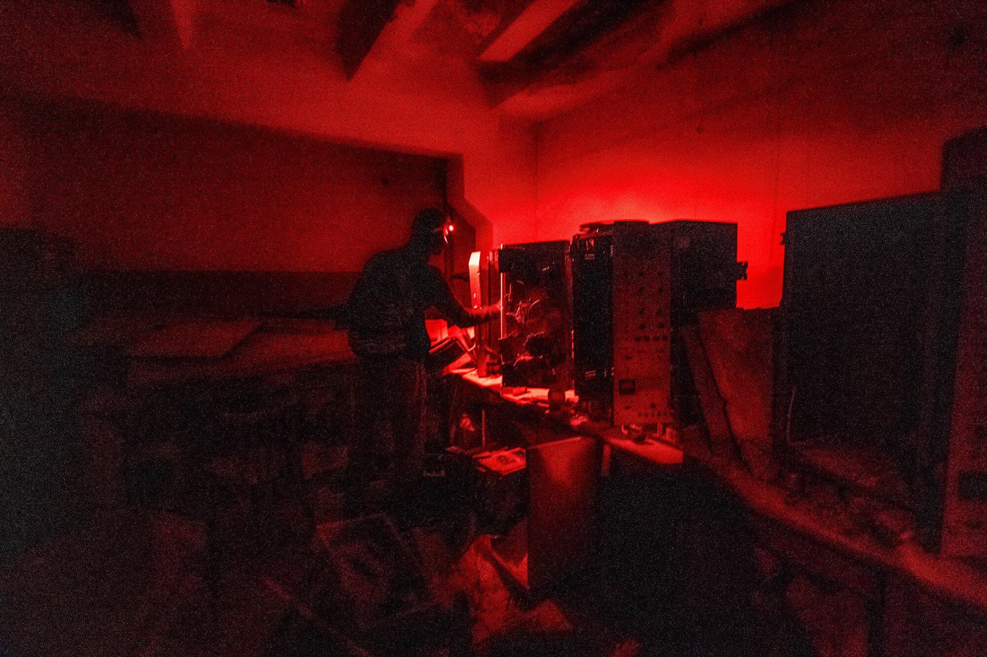 A person wearing a headlamp stands near old electronic equipment in a dimly lit room with red lighting, surrounded by cluttered objects and shadows.