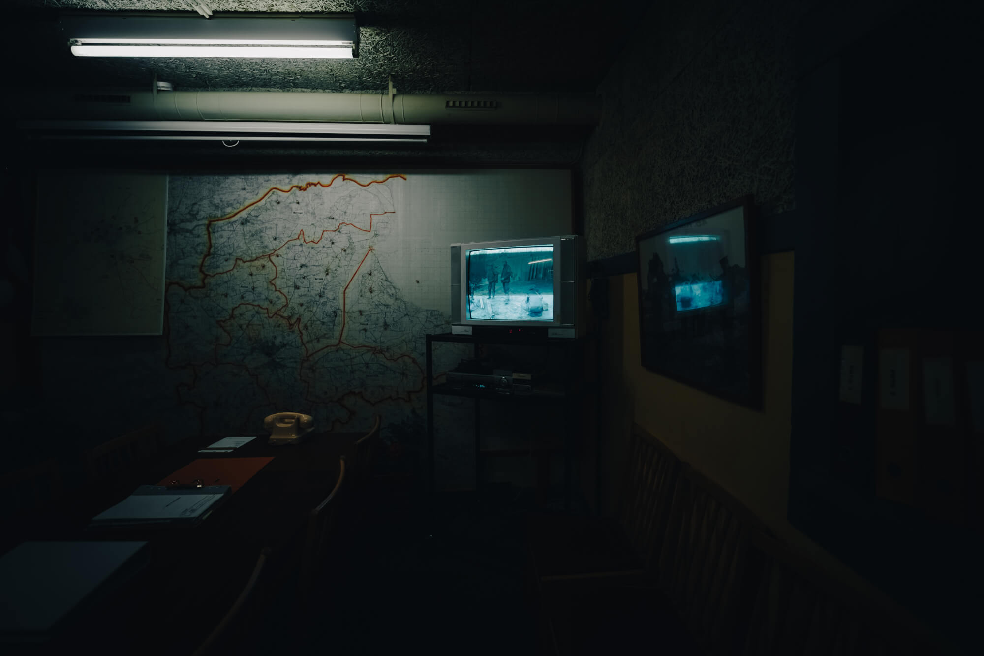A dark room with a wall map featuring orange outlines, a vintage television displaying a grainy image, a long wooden table with stacked papers, and a yellow telephone.