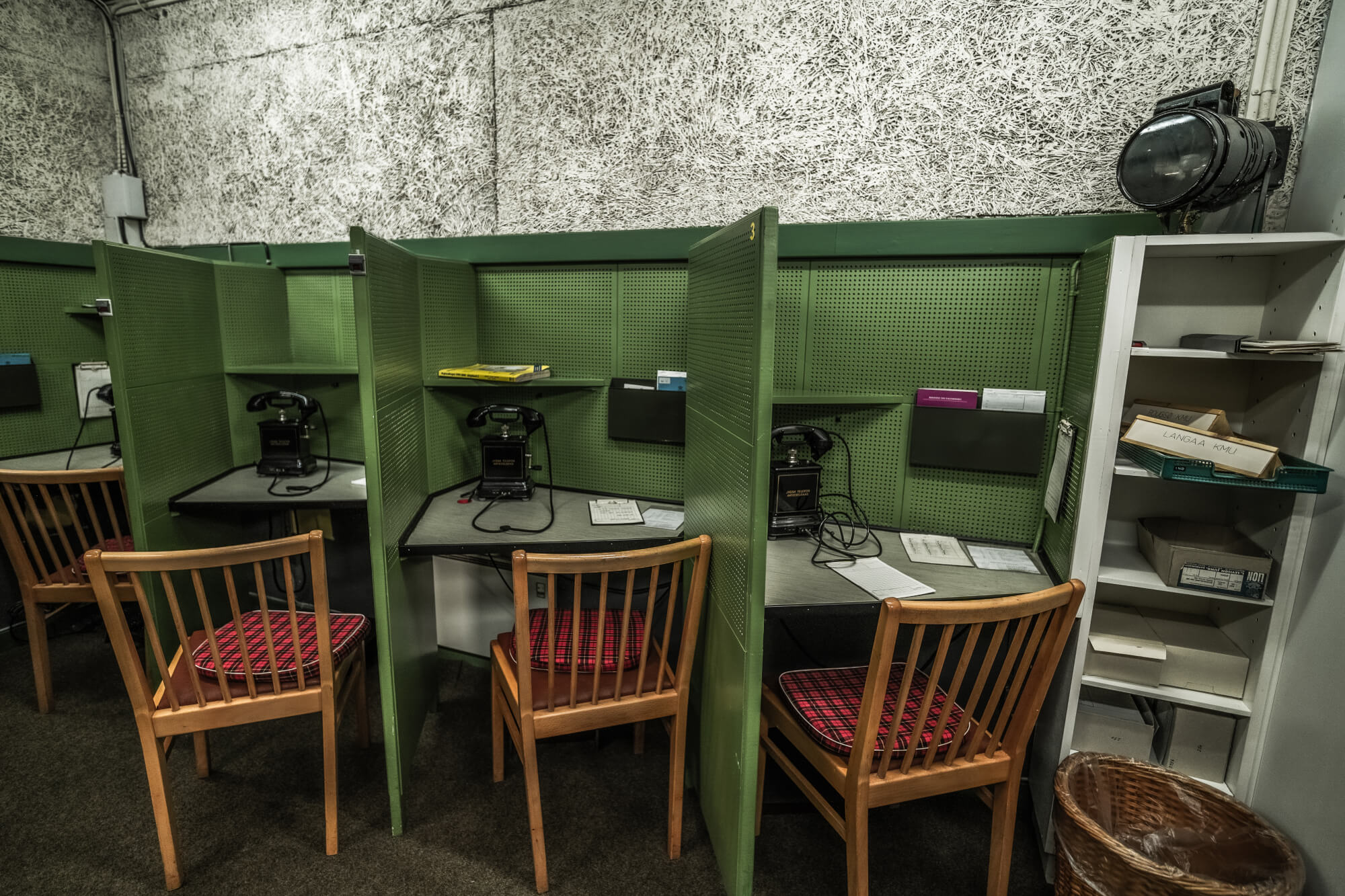 Image of vintage telephone booths with green dividers, wooden chairs with plaid cushions, rotary phones, and scattered papers on desks, set against a textured wall.