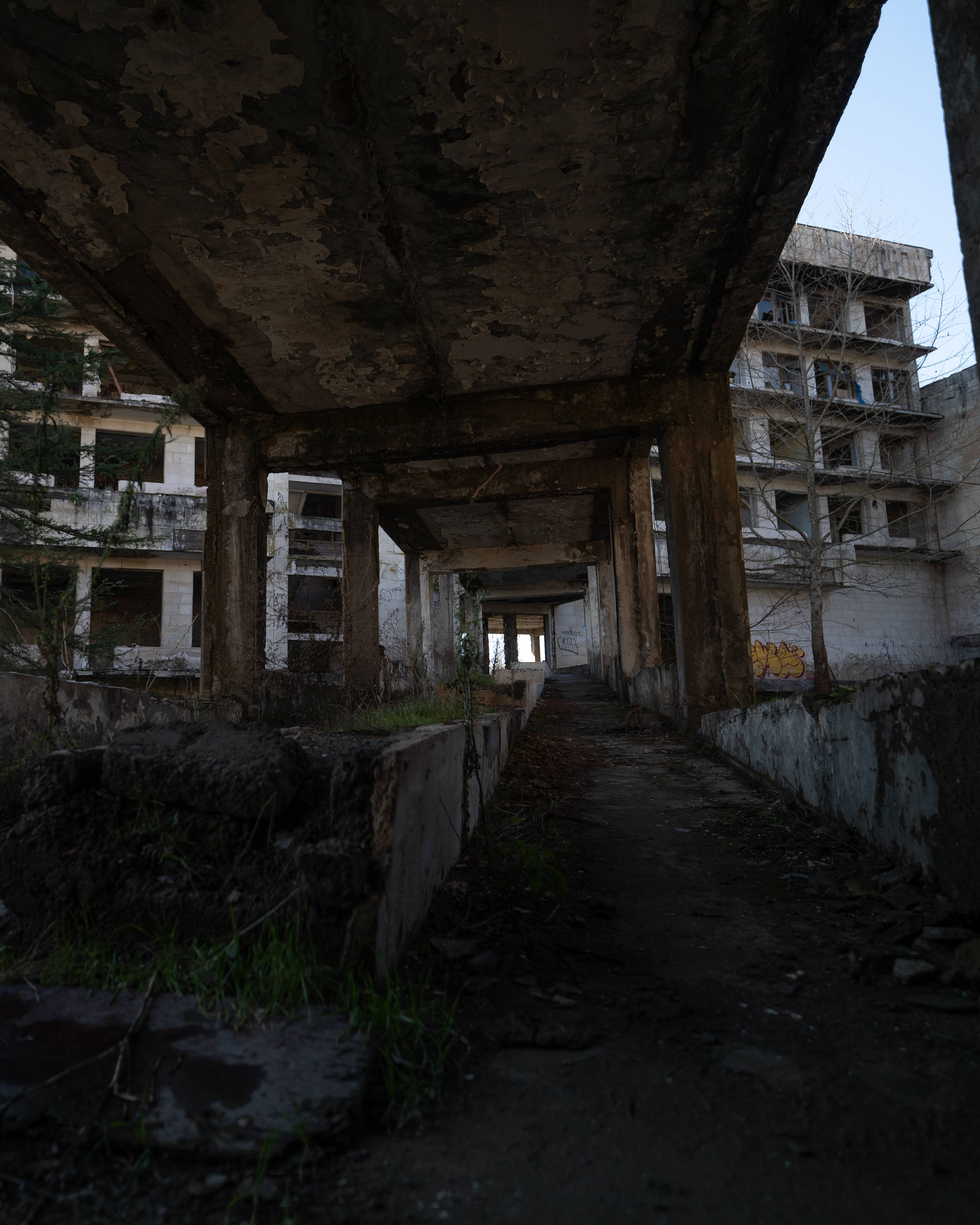 A view looking down an abandoned pathway surrounded by crumbling concrete pillars, with signs of overgrowth and neglected structures in the background.
