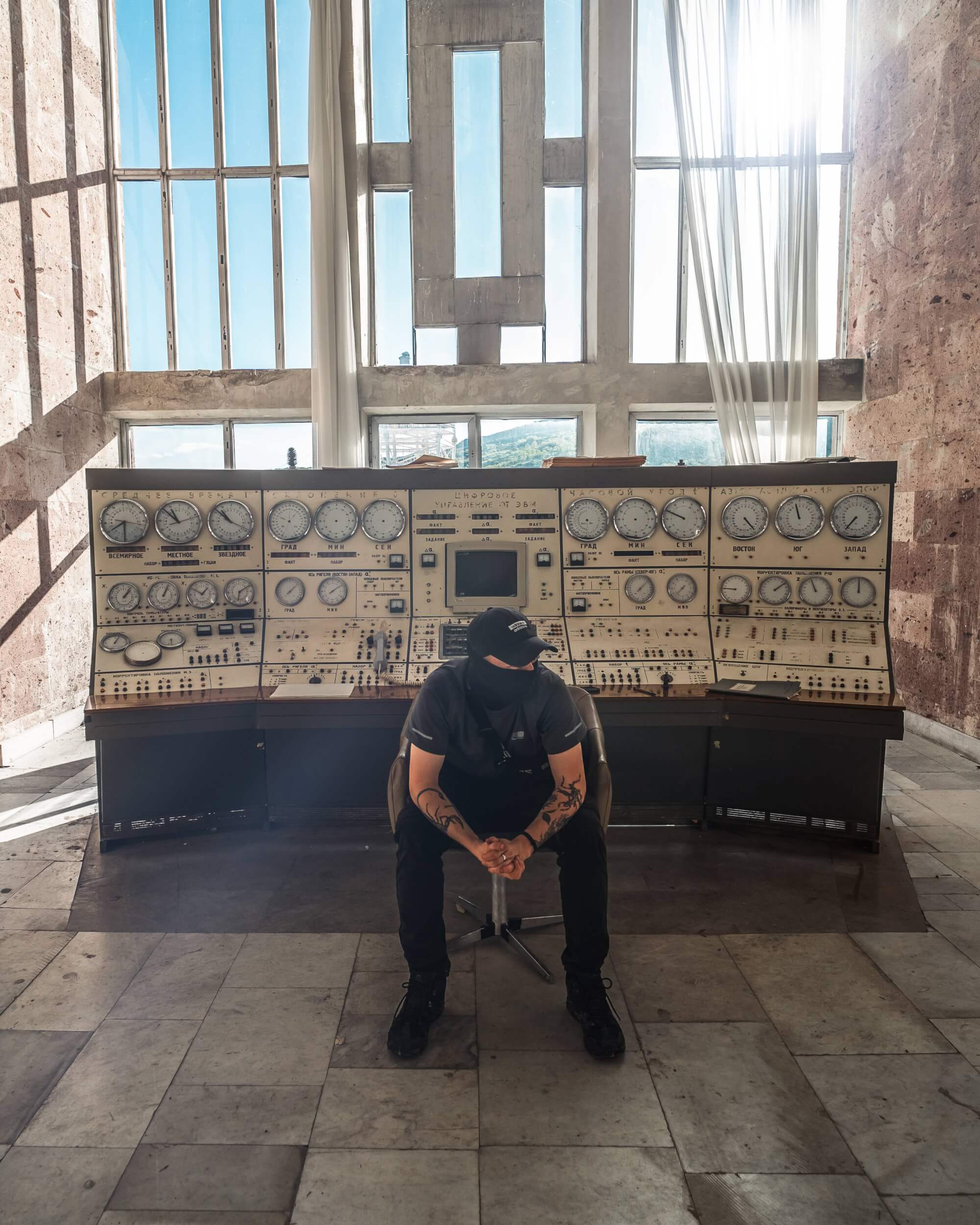 A person sits in a chair in front of a large vintage control panel in a sunlit room with large windows, casting shadows on the stone floor.