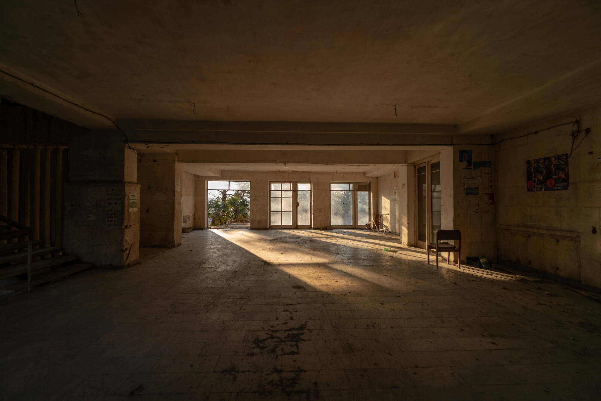 Interior of an abandoned building with large windows; sunlight casting shadows on the floor, a lonely chair in the corner, and faint greenery visible outside.
