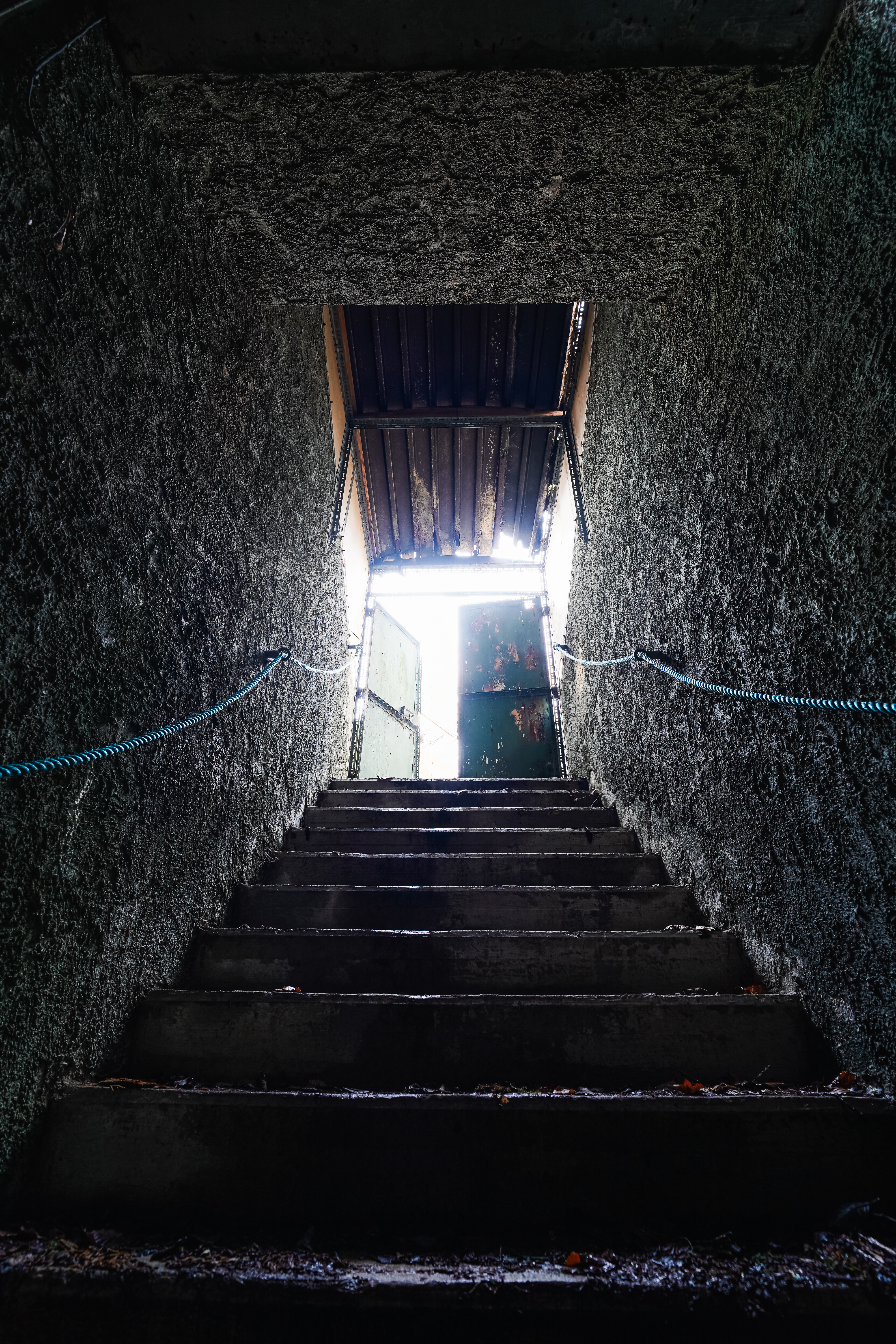 Looking up a dark stairway toward a partially open green door letting in bright light. The walls are rough and dark, with a blue rope along the side.