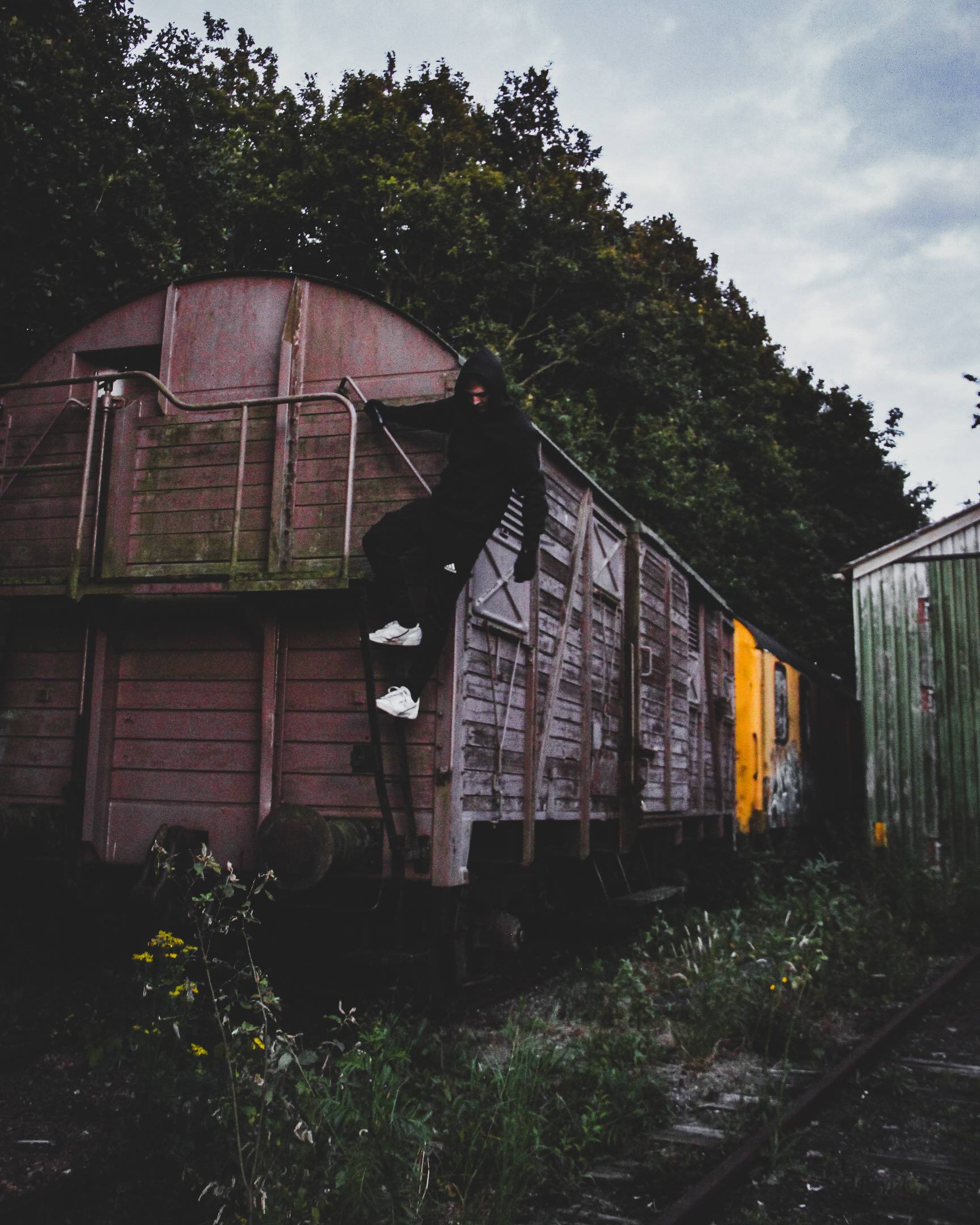 A person in a black outfit climbing down from an old rusted train car, surrounded by overgrown grass and trees under a cloudy sky.