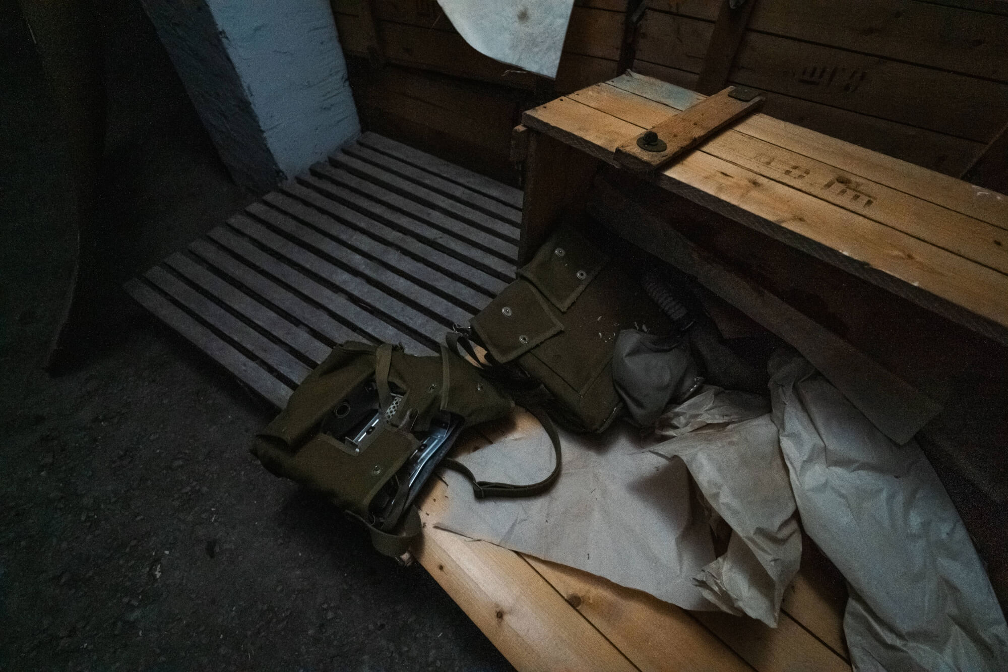 A dark corner with wooden slats on the floor, featuring two olive-green bags and crumpled paper, creating a nostalgic atmosphere.