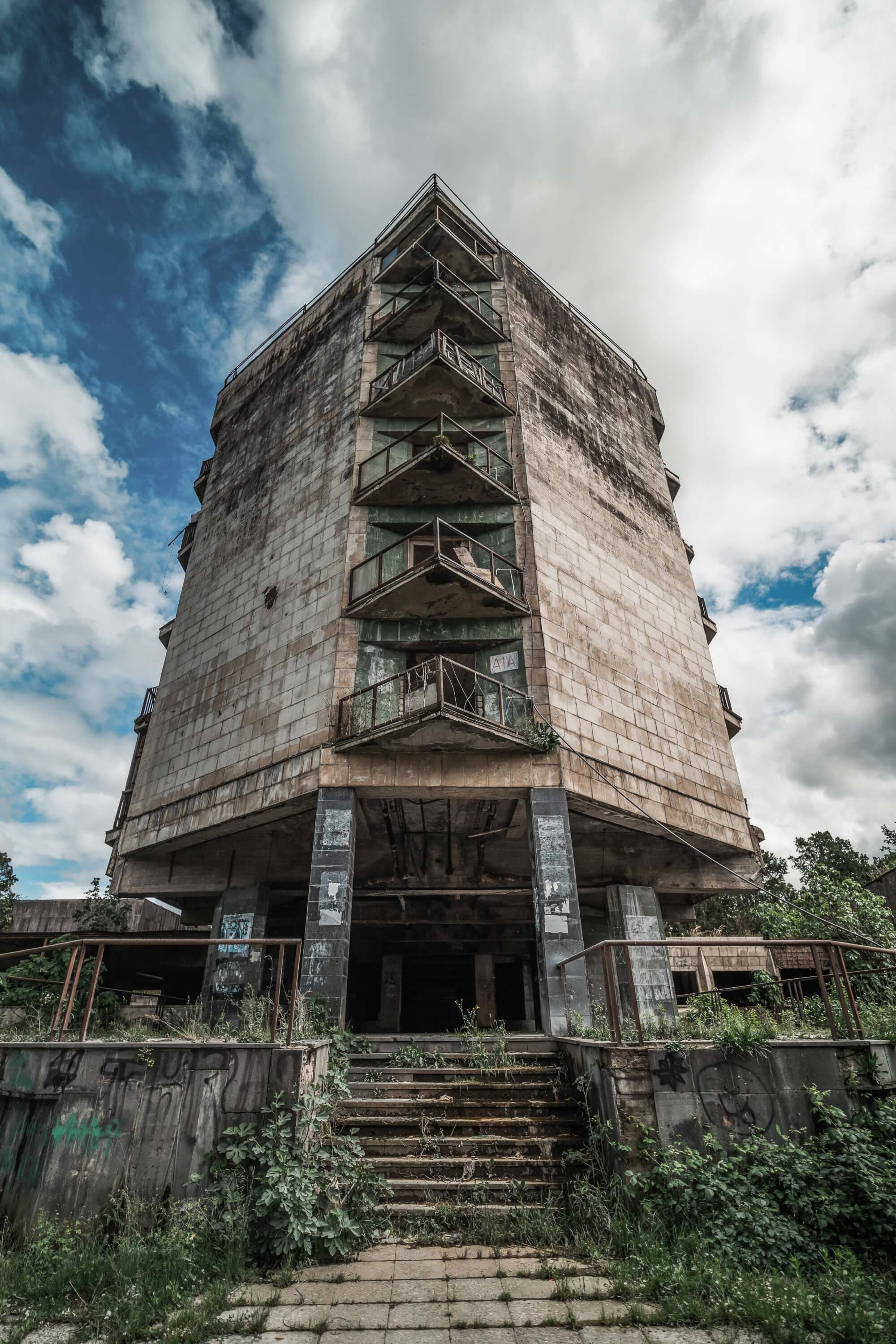 A tall, abandoned building with crumbling walls and broken balconies, surrounded by overgrown grass and plants. The sky above is cloudy with patches of blue.
