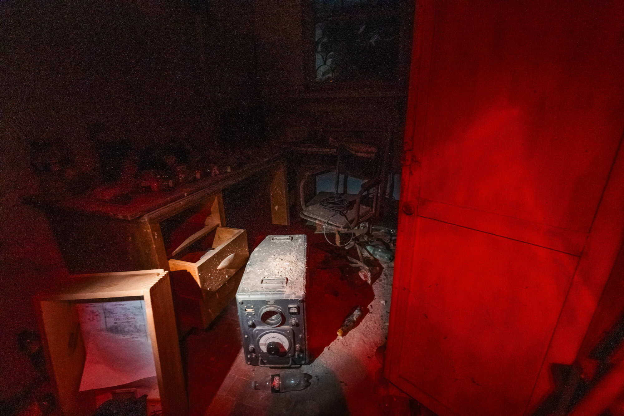 An abandoned room with a vintage radio on the floor, dust scattered around, a disheveled chair in the background, and a bright red door slightly open, creating a stark contrast with the dark and dusty surroundings.