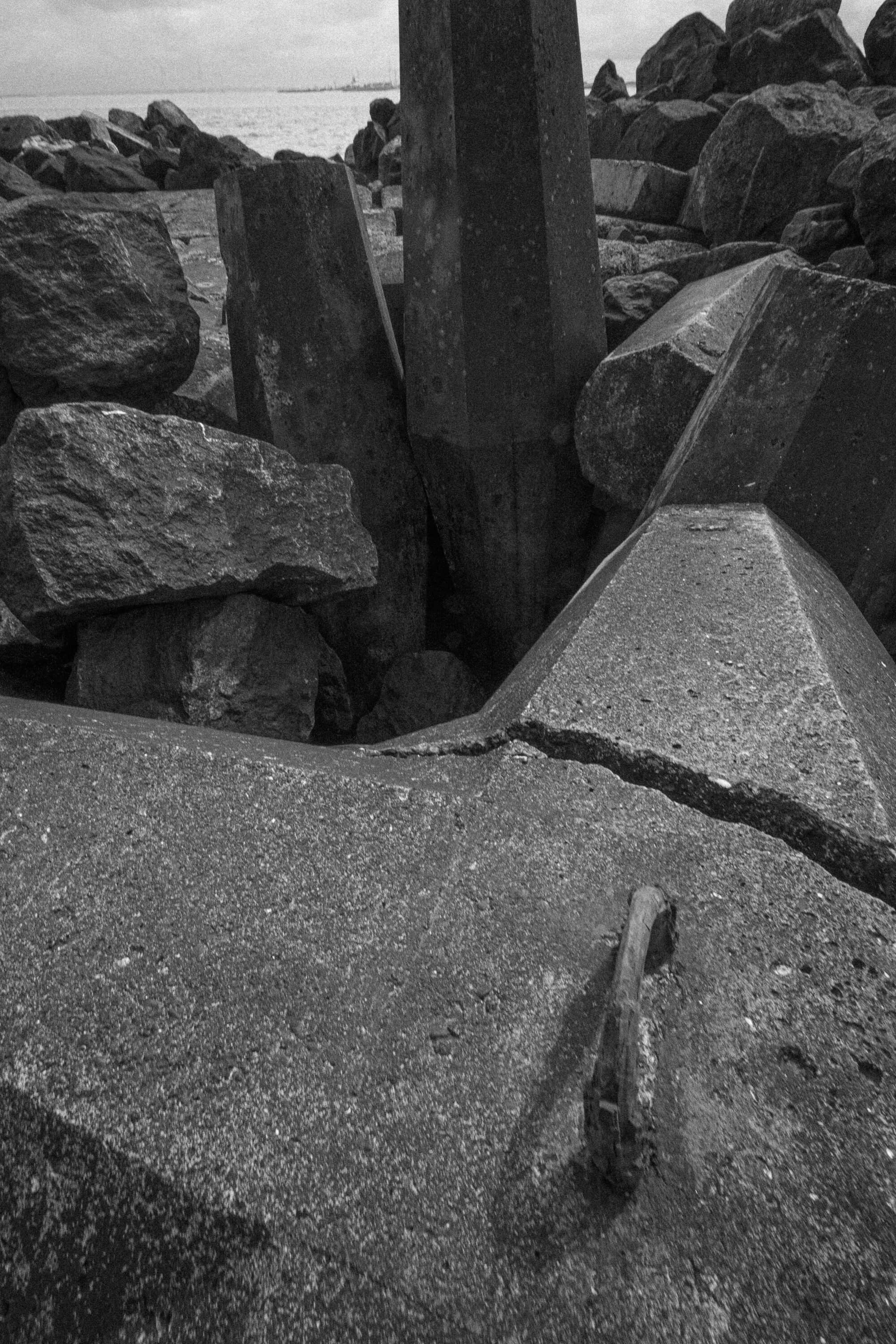 A close-up view of cracked concrete structures and large rocks by the water's edge, with a cloudy sky in the background.