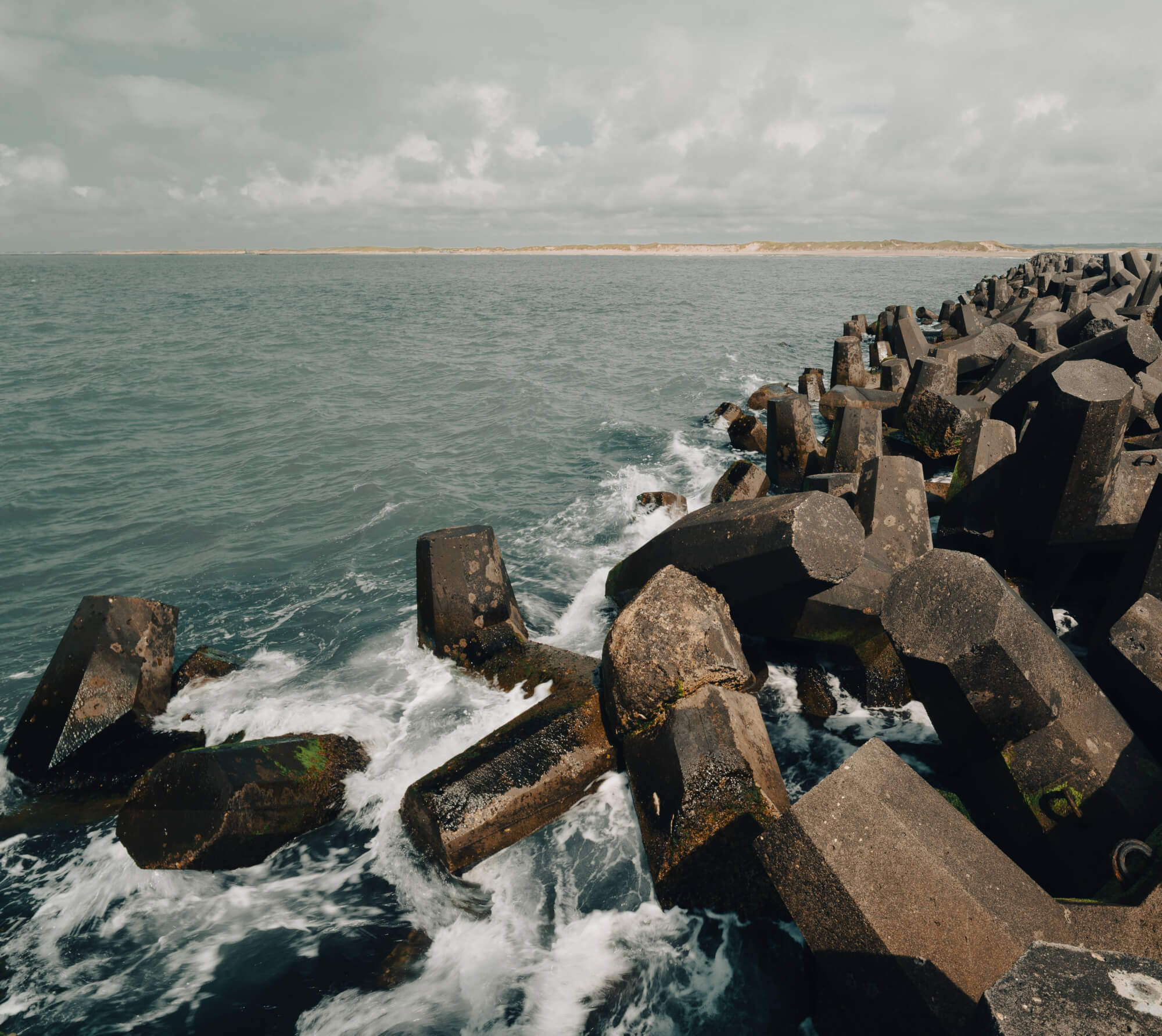 A rugged jetty made of hexagonal concrete blocks extends into a turbulent sea under a cloudy sky.