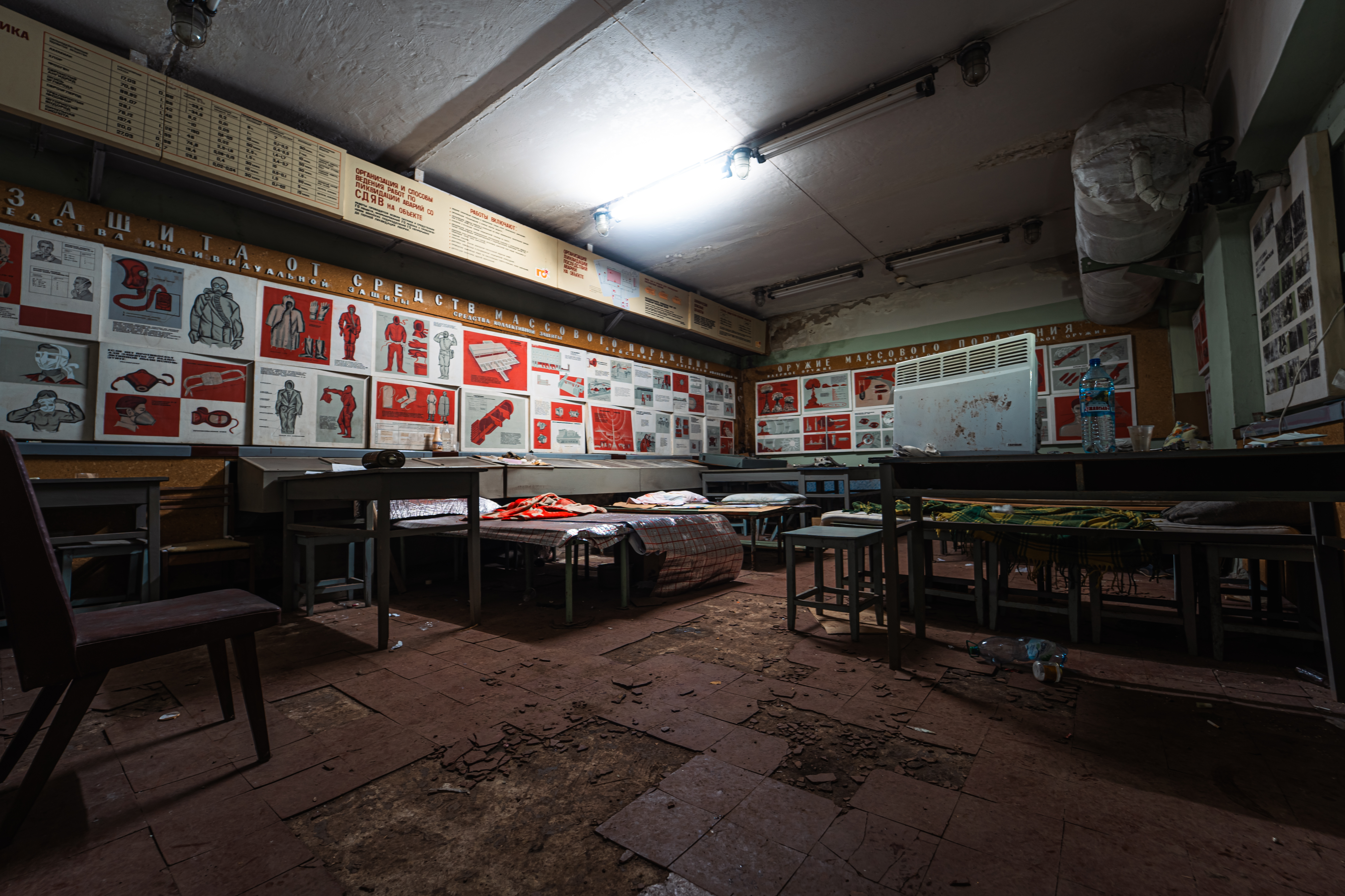 An empty, abandoned classroom with dusty floors and scattered debris. The walls are covered with red and white posters featuring Soviet-era graphics. Tables and chairs are arranged haphazardly, and a green blanket is draped over one of the tables.