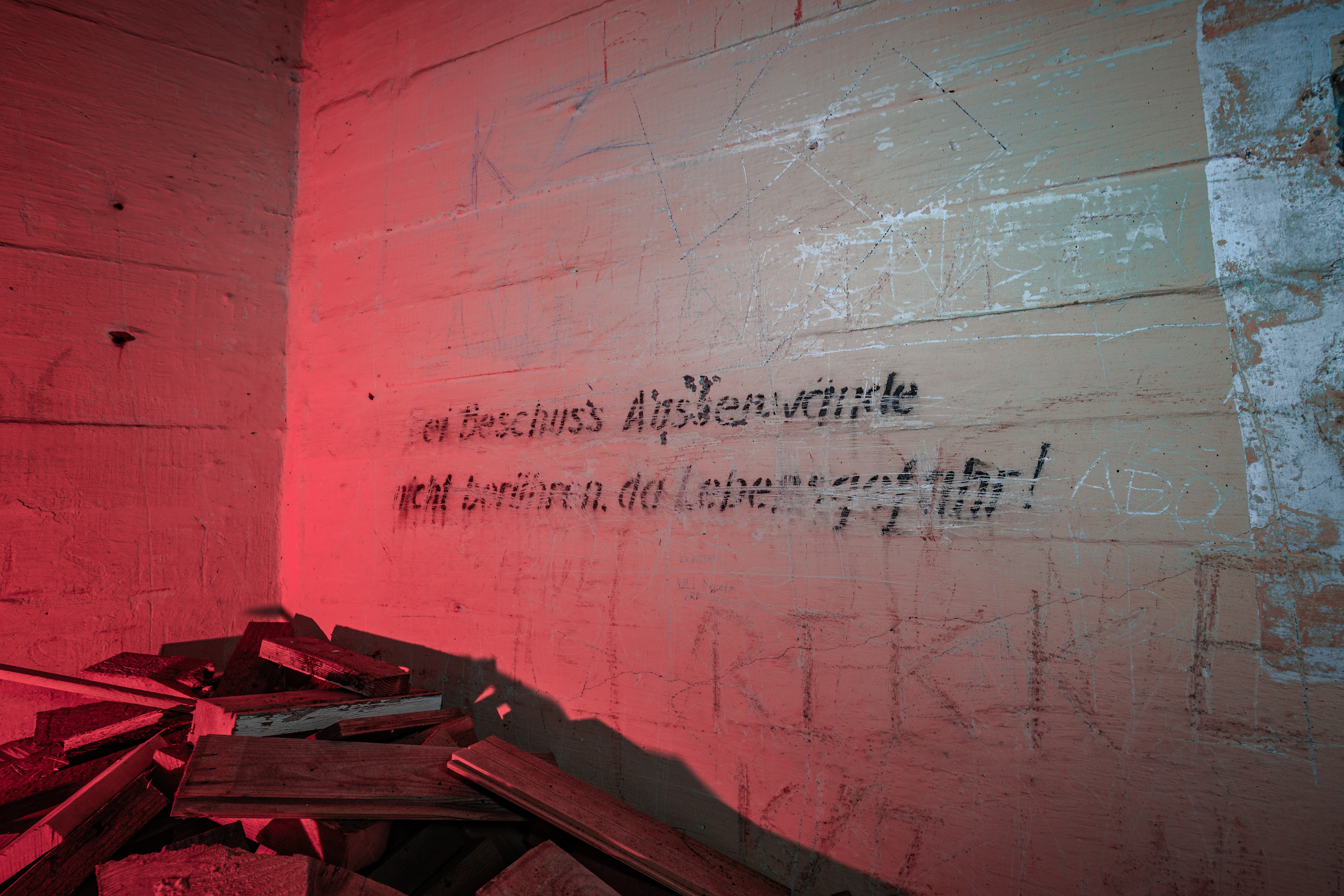 A corner of an abandoned room with wooden debris on the floor and graffiti on the wall, illuminated by a soft red light.