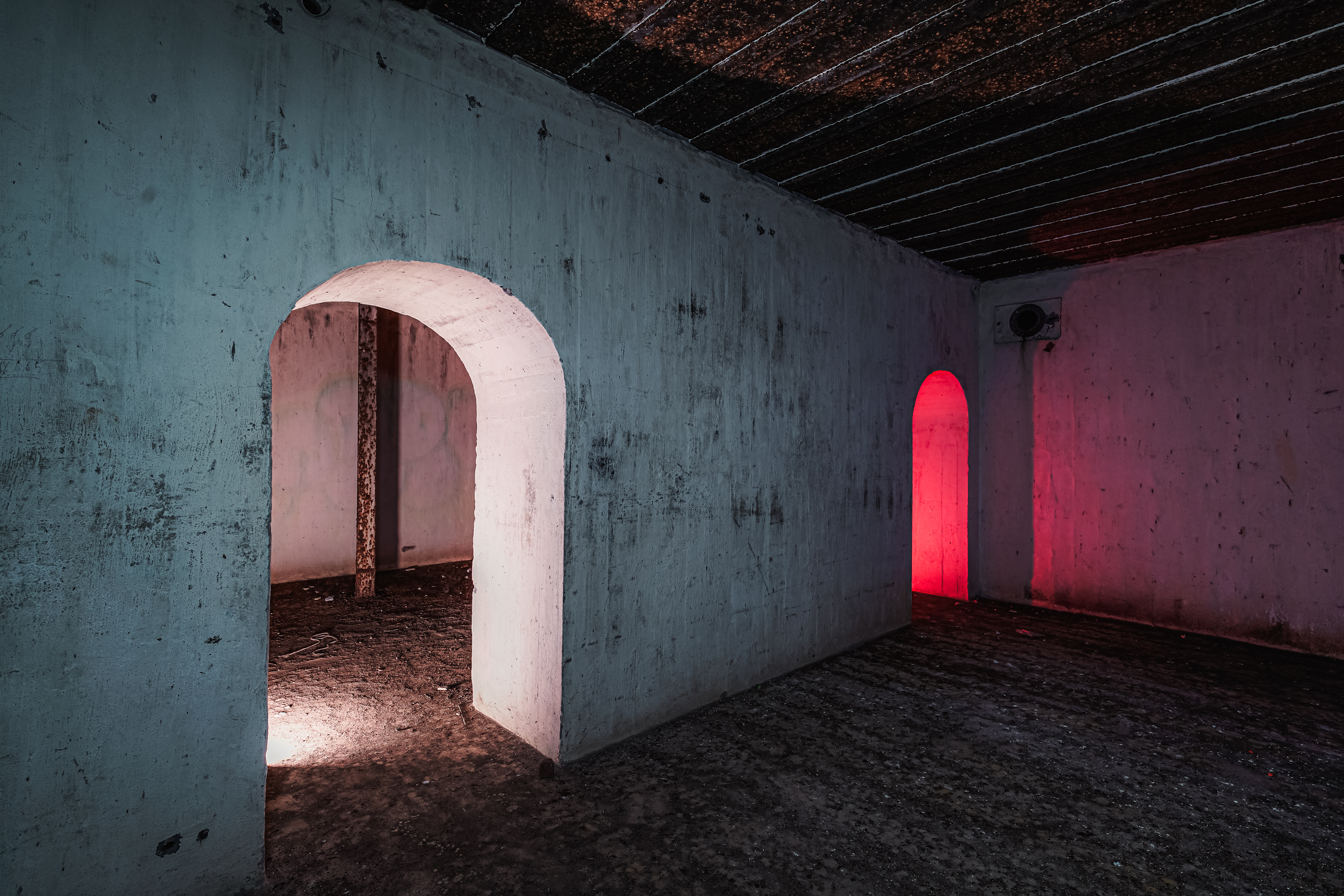 An empty room with two archways; one archway reveals a soft white light and the other a bright red light. The walls are textured and worn, contributing to an atmosphere of abandonment.