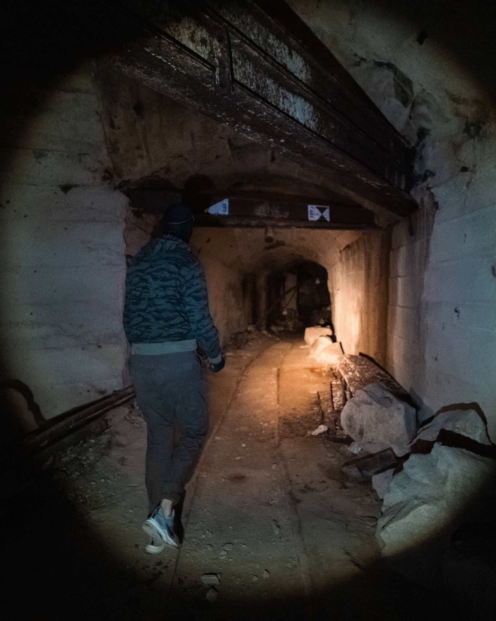 A person in a hooded jacket walking down a dark, abandoned tunnel with old railway tracks and debris on the floor.