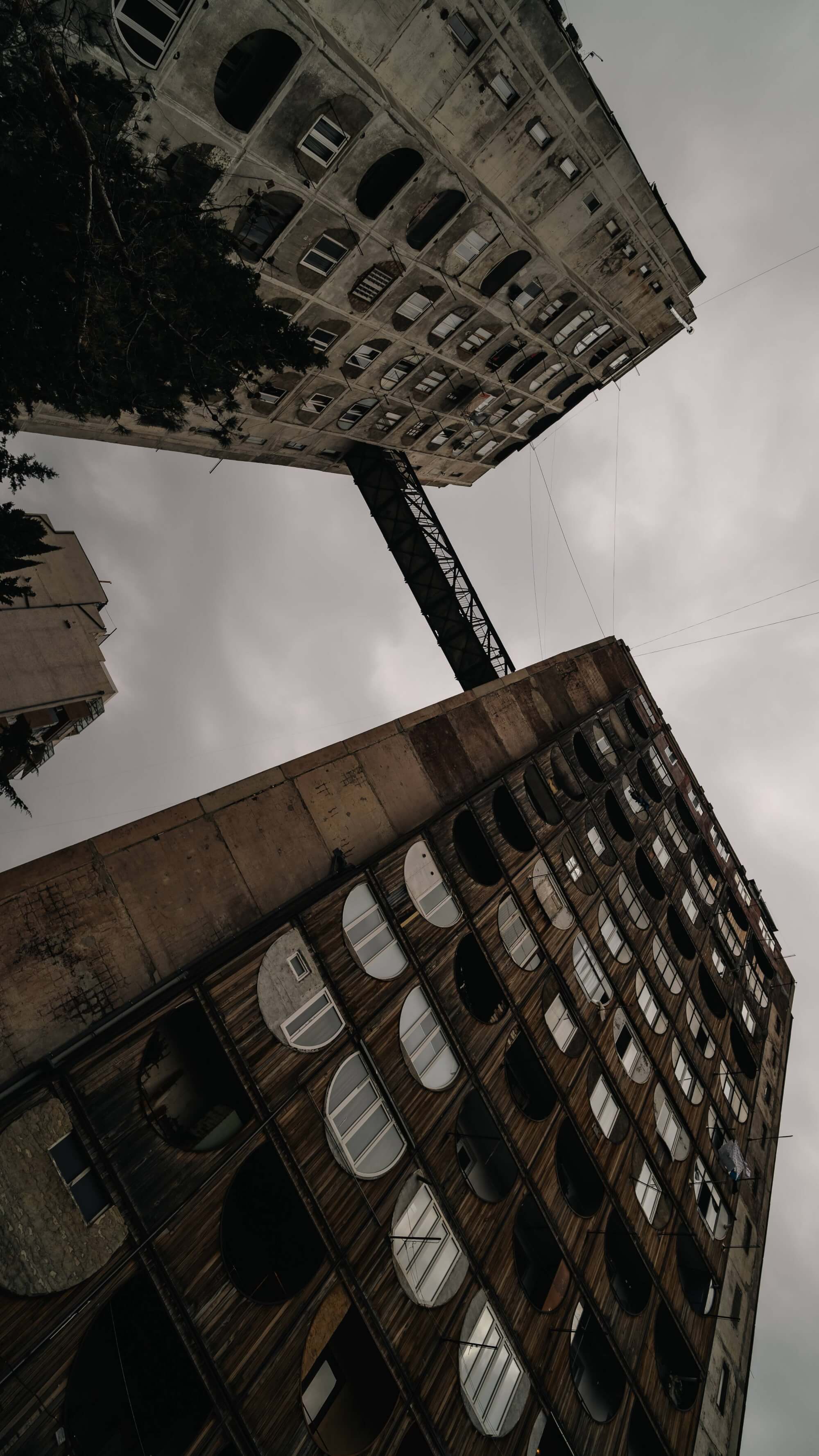 Two tall buildings: one with a wooden façade and circular windows, the other with a concrete surface. A bridge connects them above, with a gray, cloudy sky in the background.