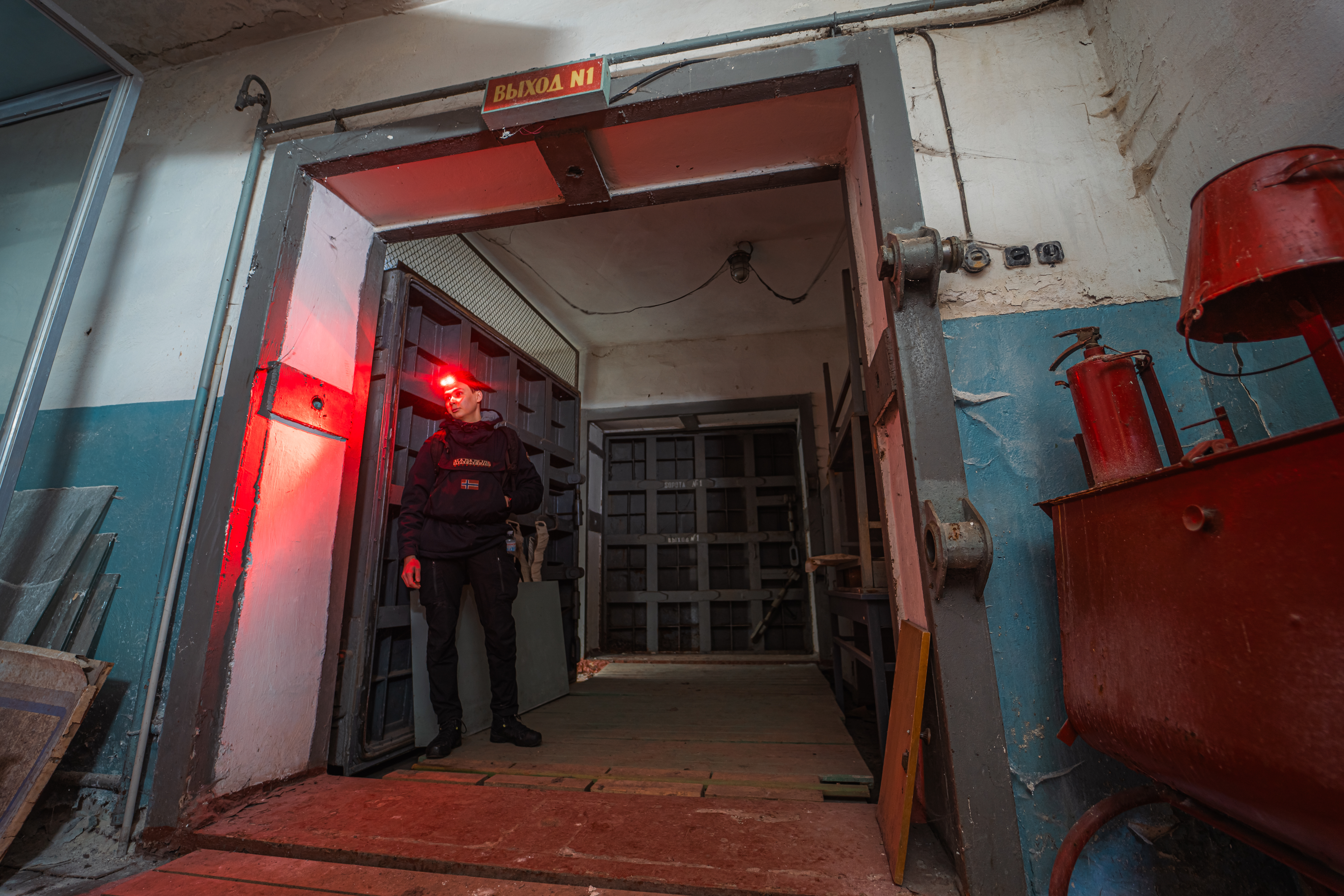A person wearing a black outfit and a red headlamp stands in a doorway of an abandoned building, with a red fire extinguisher and wooden debris visible in the foreground.